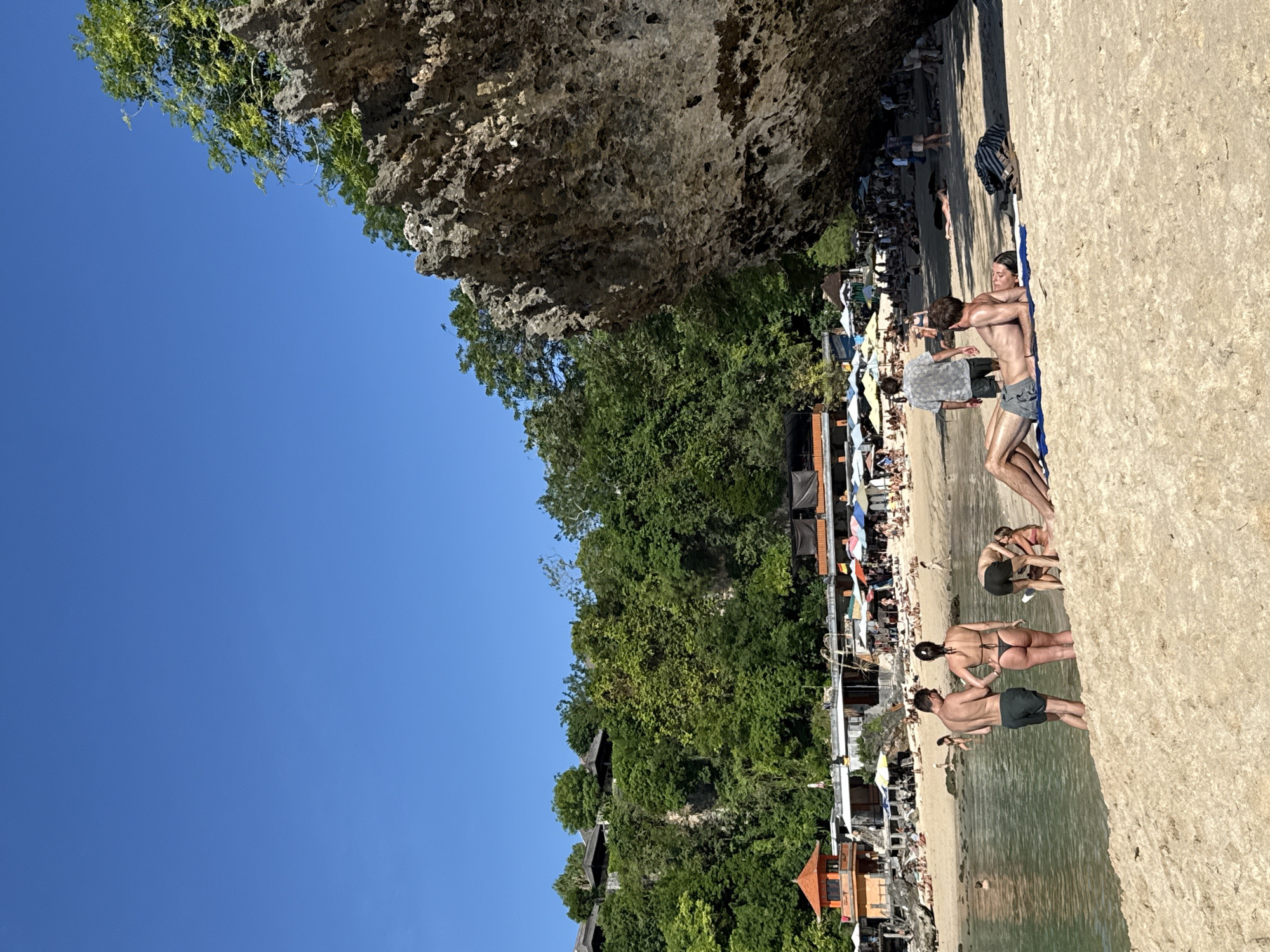 The image depicts a beach in Kuta Selatan, Indonesia. A rocky cliff overhangs the beach on the left. Trees and buildings are visible on the hillside behind the beach. On the right, people are sunbathing and swimming in the water. Some people are standing and talking near the shore, while others are lying on the sand. The sky is blue.