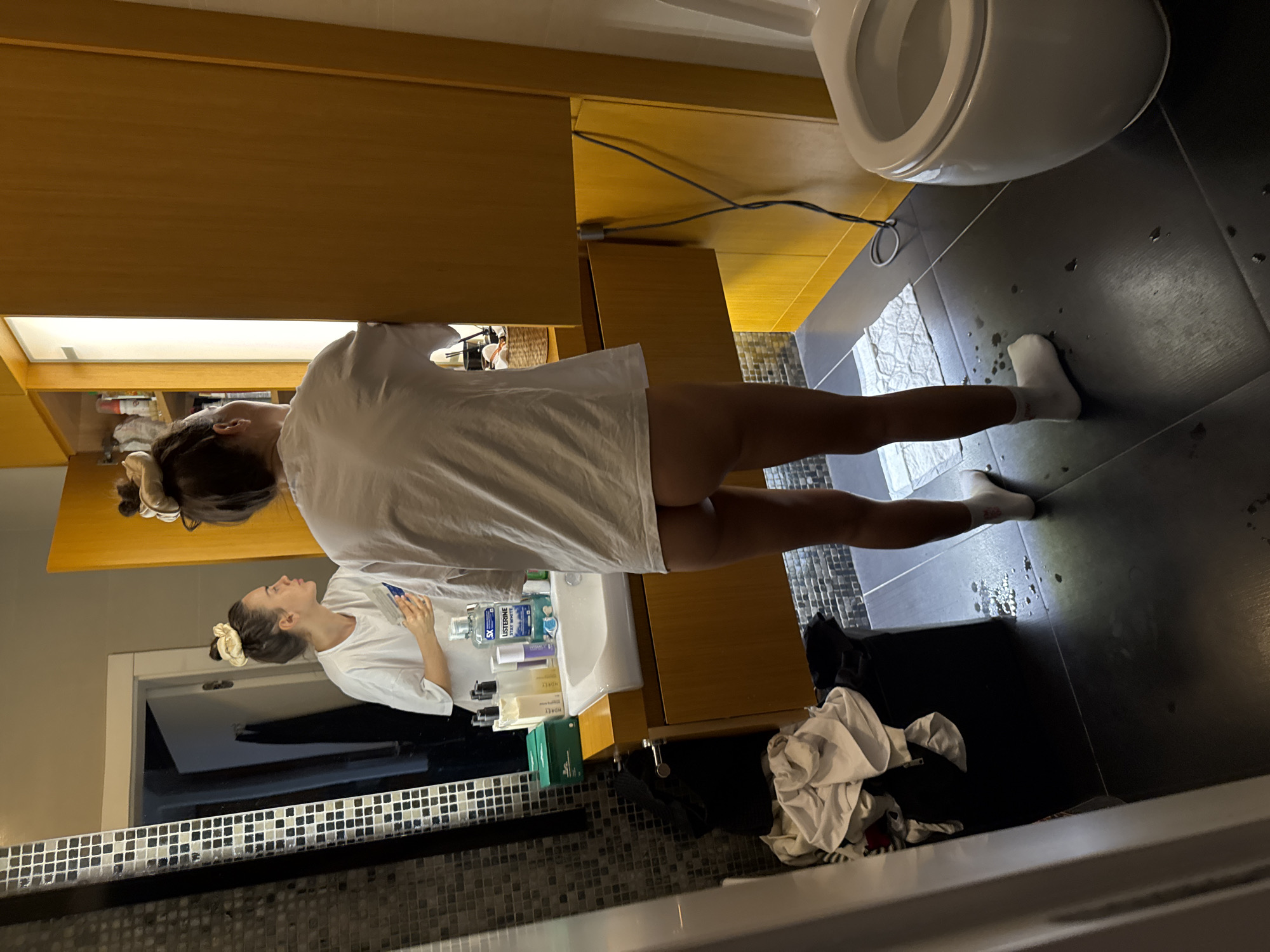 This detailed image captures a residential bathroom in Unknown, Unknown.
One individual, appearing female, is central to the image, standing with her back towards the camera, facing a mirror above a sink. She is wearing an oversized white short-sleeved t-shirt and white ankle socks. Her lower body, from the waist down to her upper thighs, is unclothed, revealing her buttocks. Her dark hair is pulled up into a loose bun secured with a light-colored scrunchie.
The bathroom features dark grey or black large square floor tiles, upon which numerous small puddles or drops of water are visible, especially between the individual's feet and extending towards the right. A small, white textured bathmat is partially visible on the floor near her right foot.
To the left of the individual, the wall is covered with a dark tile pattern featuring small, square white mosaic accents. A large rectangular mirror reflects the individual's front, showing her face in profile looking towards the right. In the reflection, she appears to be holding a white-handled tool or brush in her right hand near her face, possibly engaged in a personal hygiene routine.
Below the mirror is a wooden vanity counter and cabinet in a light brown or honey finish. A white rectangular ceramic sink with a chrome single-lever faucet is inset into the counter. Various personal care products are arranged on the counter, including a green box, multiple bottles (some black, some clear with purple contents), and a prominent blue bottle of Listerine Cool Mint mouthwash.
To the left of the vanity, a dark bin or laundry basket is overflowing with discarded white and dark clothing items.
To the right of the individual, a tall, floor-to-ceiling wooden cabinet structure, matching the vanity, is visible. An electrical cord, dark in color, extends down the right-most wooden panel. In the lower right of the frame, the front portion of a white ceramic toilet with its lid closed is visible.