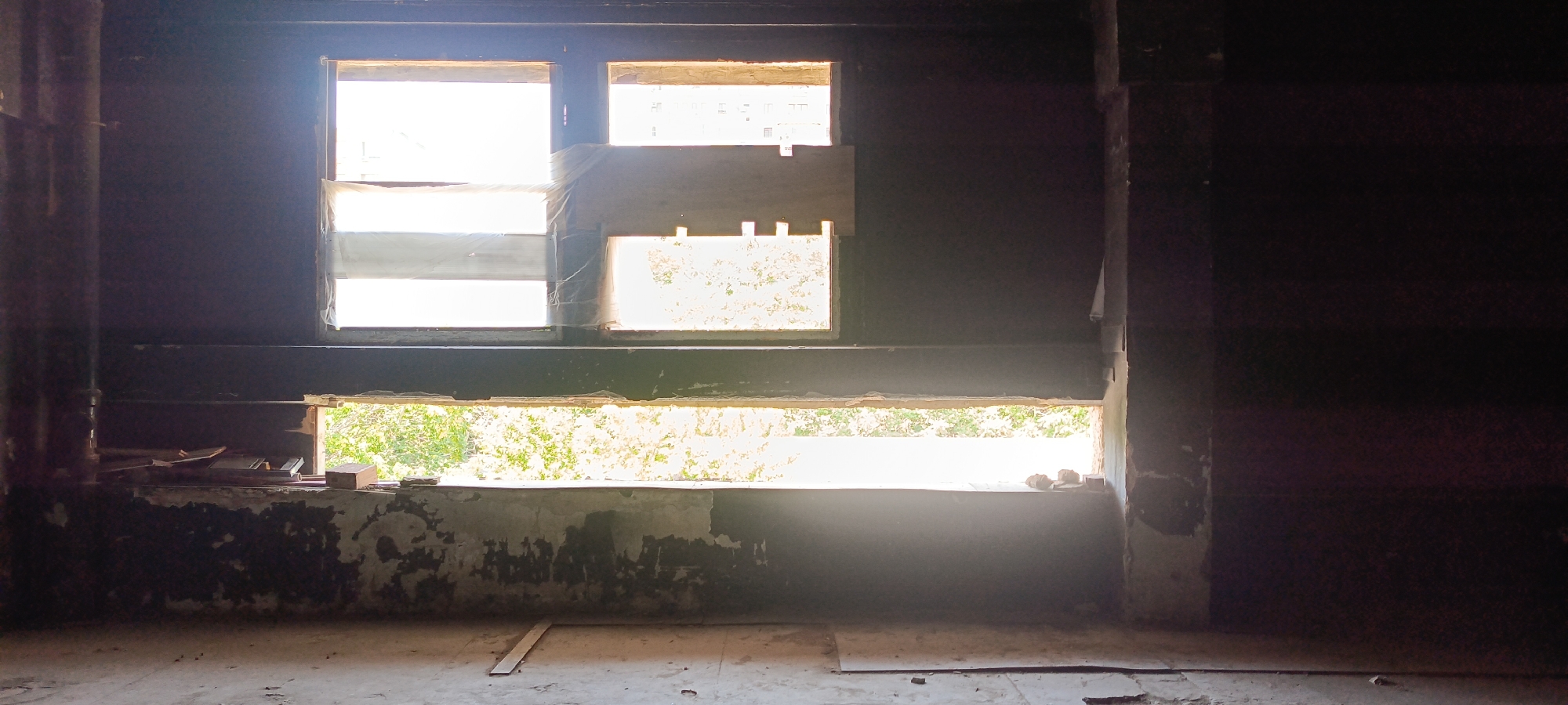 The image depicts the interior of a dark, unfinished room with several windows allowing natural light to enter. The windows are of varying sizes and shapes, with some covered by white plastic sheeting. Through the windows, glimpses of greenery and buildings can be seen. The floor is concrete and littered with debris, and the walls appear to be rough and unpainted. The overall impression is one of abandonment or ongoing construction. The scene is located in Baku, Azerbaijan, though the specific building or area is not identified.