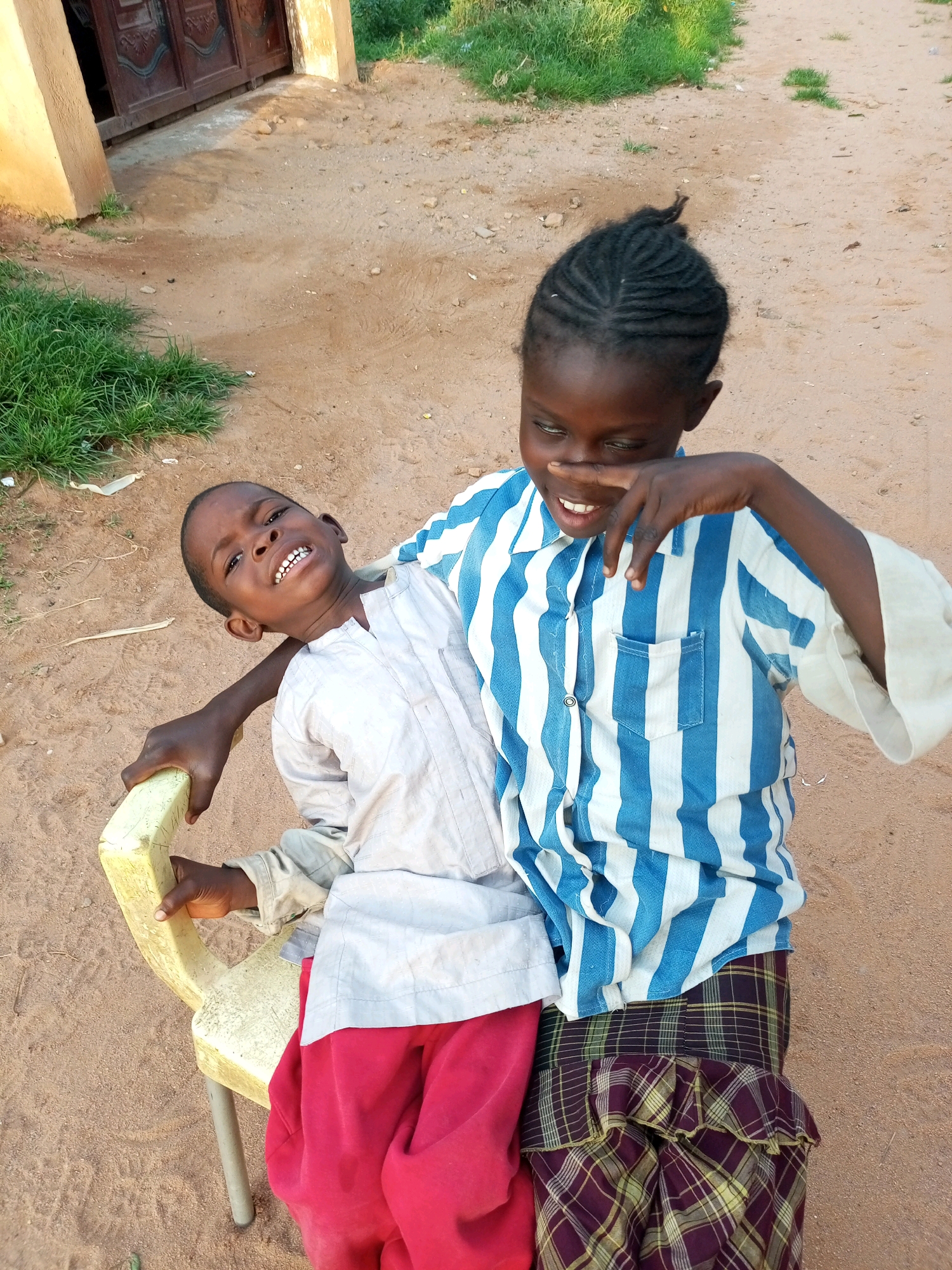 Two children are sitting on a yellow plastic chair outdoors in Tumfure, Nigeria. The child on the left, a boy, is looking up and smiling widely, showing his teeth. He is wearing a light-colored, long-sleeved shirt and red trousers. The child on the right, a girl, is leaning over and touching her nose with her finger, looking down with a playful expression. She has braided hair and is wearing a blue and white striped shirt and a plaid skirt. The ground is sandy and dry, with some dry grass and a building with large wooden doors visible in the background. The scene appears to be in the late afternoon or early evening, suggested by the warm lighting.