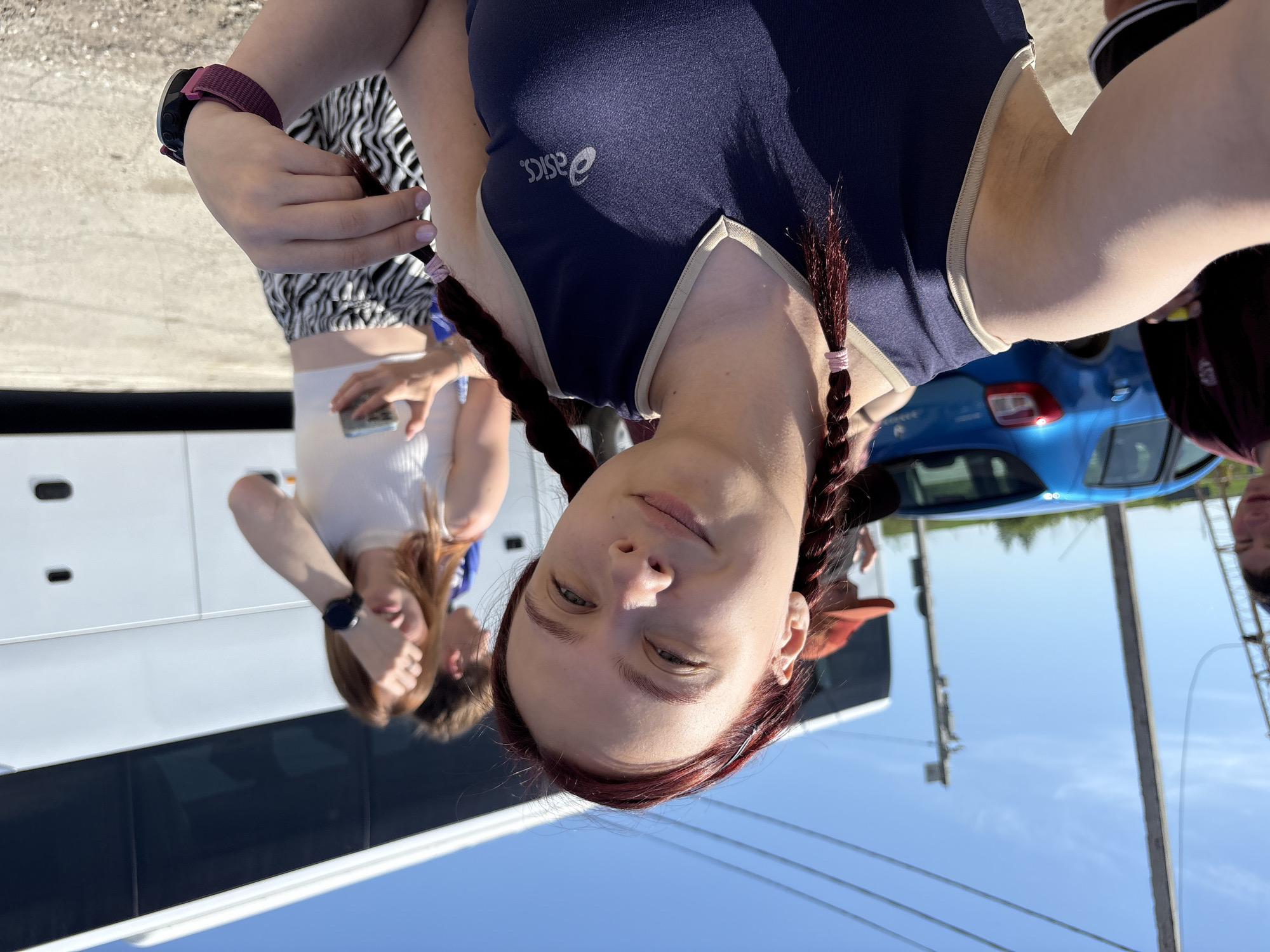 The image shows a woman with red hair braided into pigtails, wearing a navy blue and white Asics-branded athletic outfit. She is standing outside, with a light blue sky in the background. A white bus is visible on the left, and a light blue car is behind her. Other people are also present, including a woman in a white shirt and zebra-print shorts holding a cell phone, and another person partially visible on the right. The location is identified as Grishanki, Russia, based on the task request.