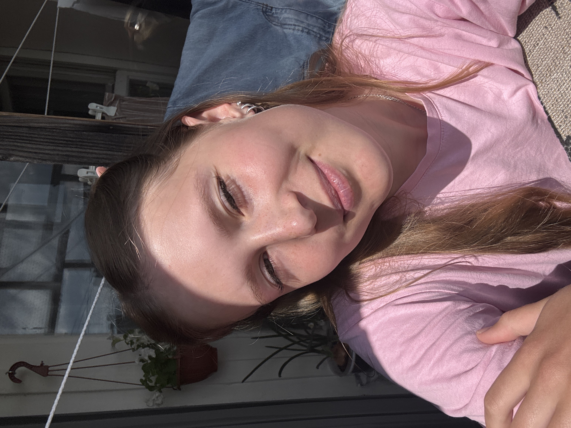 The image depicts a close-up portrait of a young female in an outdoor setting, likely a balcony or terrace in Chaikovsky, Russia. The subject, a fair-skinned individual with light brown hair exhibiting auburn undertones, is positioned centrally, looking directly at the camera with a gentle smile. Her eyes are slightly narrowed, a probable reaction to the bright sunlight illuminating the scene from the left, casting distinct shadows across the right side of her face and torso. Freckles are visible on her nose and cheeks. She wears a plain, light pink, short-sleeved t-shirt and a thin silver chain necklace. A small, spiraled silver hoop earring is visible in her right ear (viewer's left).
In the background, behind the subject's left shoulder (viewer's right), a segment of a vertical wooden post or beam is visible. To its right, a blue denim jacket hangs on a white clothesline, secured by white clothes pegs. Behind this, a window or paneled structure reflects bright light.
To the subject's right (viewer's left), a dark grey vertical structure borders a lighter wall. A red, '2'-shaped hook supports a terracotta-colored hanging pot containing white-flowered plants. Below this, a cylindrical pot holds a green, spiky plant, possibly a succulent. A thin white line, consistent with a clothesline, extends horizontally across the upper left quadrant of the background. The bottom right corner of the image shows a textured, light brown woven surface, possibly a mat or rug. The subject's left hand (viewer's right) is partially visible in the bottom left.