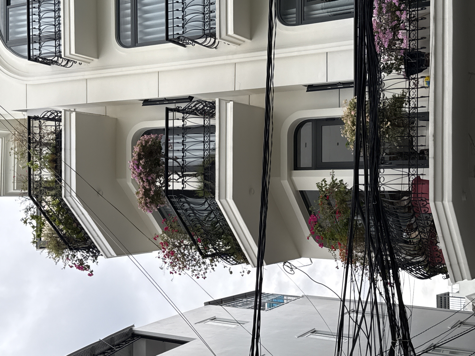 A low-angle exterior shot captures the side of a white multi-story building in Nha Trang, Vietnam, with overcast skies overhead. The building features balconies with black metal railings adorned with plants and flowers. On the balconies, potted plants with pink and purple blooms are visible. The building has arched windows with dark frames and curtains. Adjacent to this building, on the left, is another white building with several windows and a metal grate structure. A dense network of black electrical wires spans across the foreground, obscuring parts of the buildings and balconies. The scene appears to be in an urban area, likely a residential or commercial district.