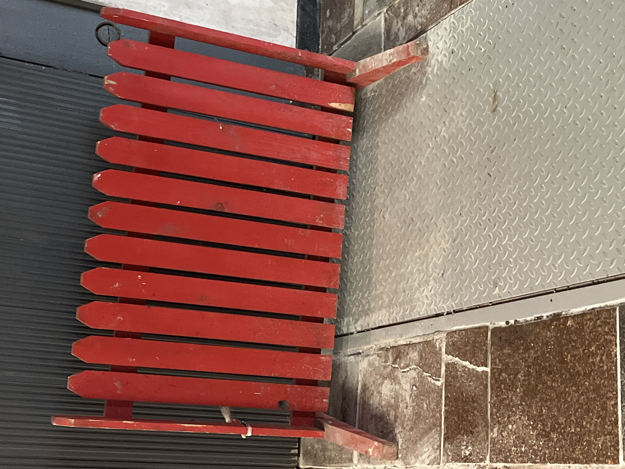 A freestanding, weathered red wooden barrier, constructed from vertical pickets with pointed tops and supported by two end posts with outward-extending feet, is positioned across a floor transition. The barrier's red paint is chipped and scuffed, exposing lighter wood in numerous areas. Its left base rests on cracked, brown square tiles, while its right base is situated on a light-gray metal diamond plate surface, separated by a thin metal strip. Behind the barrier, a dark, vertically corrugated surface, consistent with a roll-down shutter or wall panel, dominates the background. A small white tie is loosely affixed to the left vertical support post of the barrier. The image contains no visual cues, such as signage, landmarks, or unique architectural features, to determine a specific geographic location, including the city of Minsk, Belarus. No people or interactions are depicted.