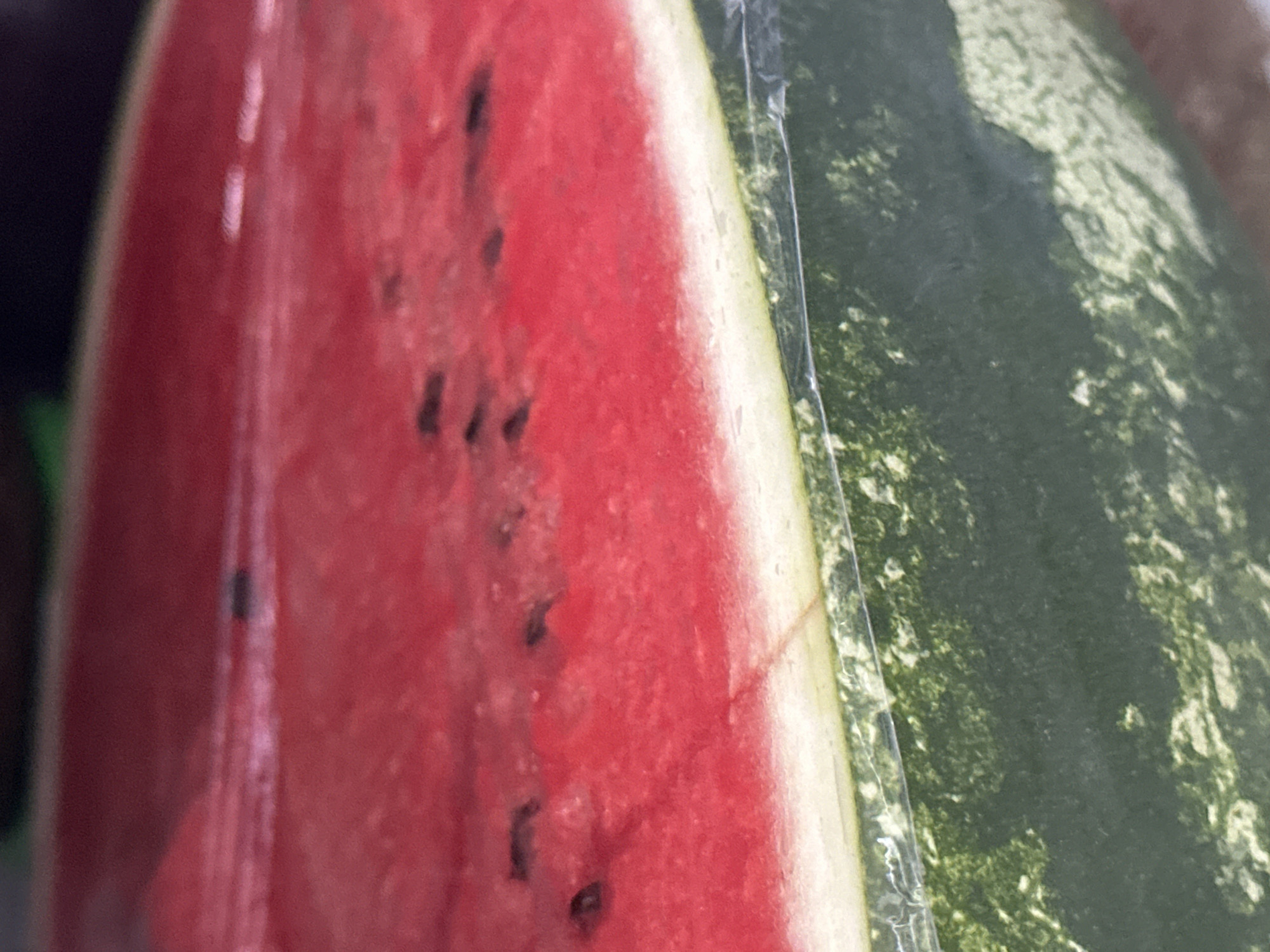 A close-up shot of a cut watermelon shows the bright red flesh speckled with black seeds. The rind, with its characteristic green stripes and lighter mottling, is visible at the bottom. The fruit appears to be wrapped in clear plastic. The scene is indoors, likely a market or grocery store, as suggested by the indistinct background and the presence of produce. This image was taken in Nevinnomyssk, Russia.