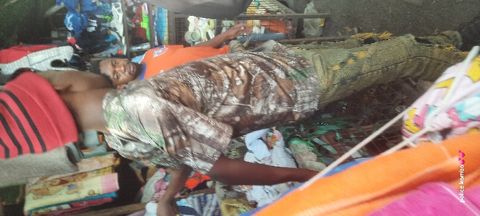 Two men are seen reclining outdoors. One is partially shirtless, wearing a red towel with black stripes, and is lying on a stack of folded clothes. The other man wears an orange t-shirt and camouflage pants. Both are lying down amidst piles of clothing and miscellaneous items. The setting appears cluttered and somewhat disorganized.