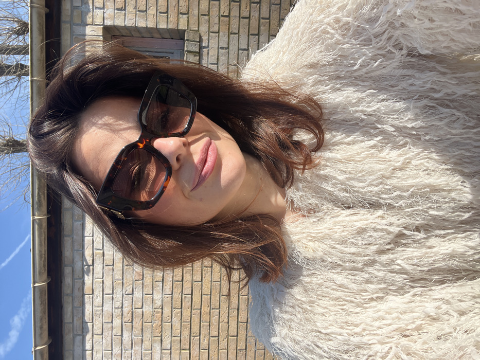 The scene depicts a female individual outdoors in Coudekerque-Branche, France. She has medium brown hair and is wearing oversized, dark tortoiseshell-patterned sunglasses with dark lenses, featuring a small golden emblem on the left temple. Her attire includes a light-colored, possibly cream or off-white, shaggy or fringed jacket. The subject exhibits a slight smile and is looking towards the camera. The background consists of a light brown brick wall, topped by a dark rain gutter. Above the gutter, bare tree branches are visible against a clear blue sky, which contains a single white contrail. The environment is brightly illuminated by direct sunlight.