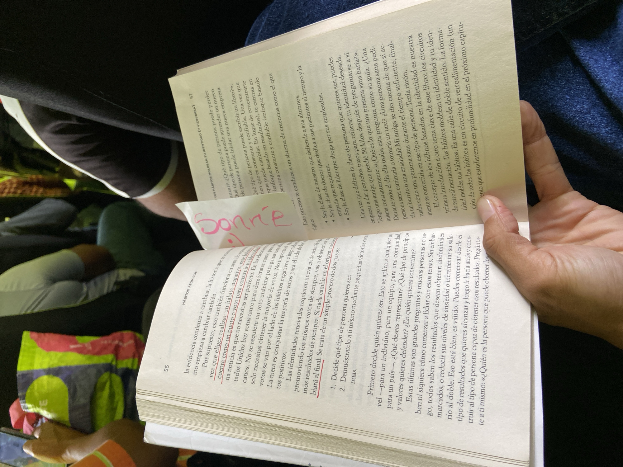 The image shows a person holding an open book. The book appears to be in Spanish and discusses habits. A handwritten note is visible on the page. The environment surrounding the person is not distinctly visible, but hints of clothing and possibly an object resembling a bag or purse are noticeable in the background. There is no information to confirm the location in Turmero, Venezuela.