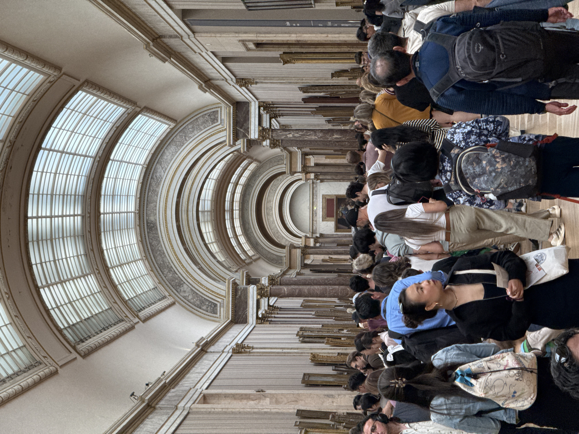 This is an interior shot of the Louvre Museum in Paris, France. A large crowd of people lines a long hallway with arched windows on the left. The walls are decorated with framed artwork. The ceiling is high and ornate, featuring intricate architectural details. People are looking at the artwork on display.