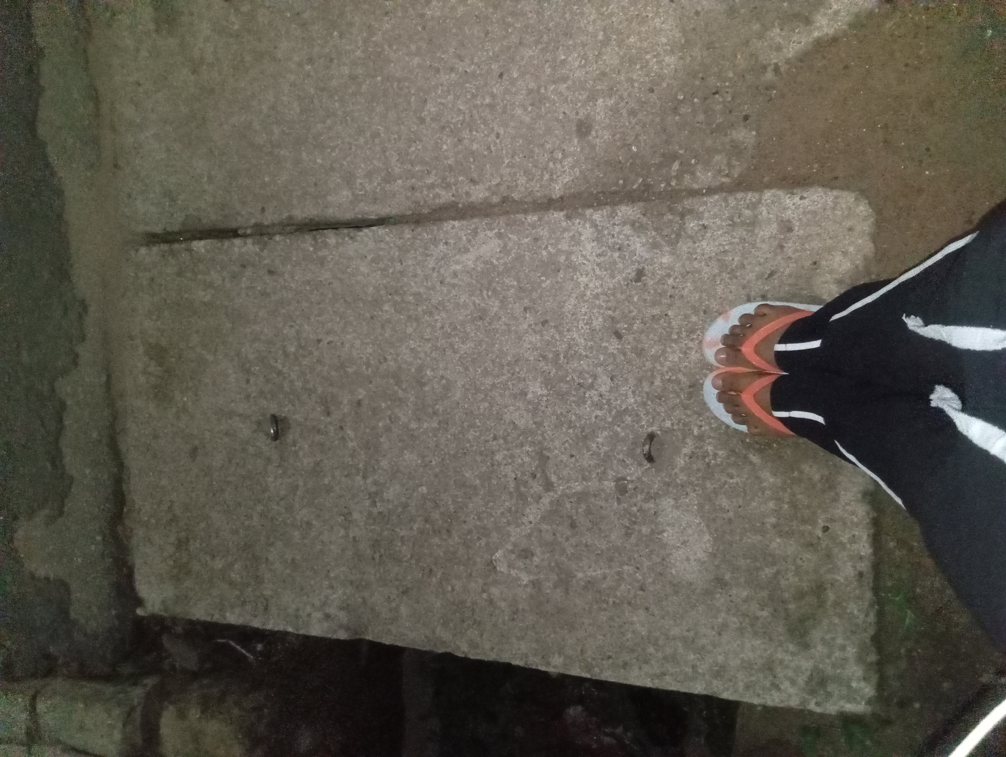 A pair of feet wearing orange and white flip-flops and black pants with white stripes stand on a concrete surface. The concrete is uneven and appears to be made of two large slabs, separated by a crack. The surrounding environment is dark and appears to be outdoors, possibly in a natural setting or an unlit urban area. The lighting is dim, casting shadows on the concrete. There are no other people or discernible objects in the immediate vicinity.