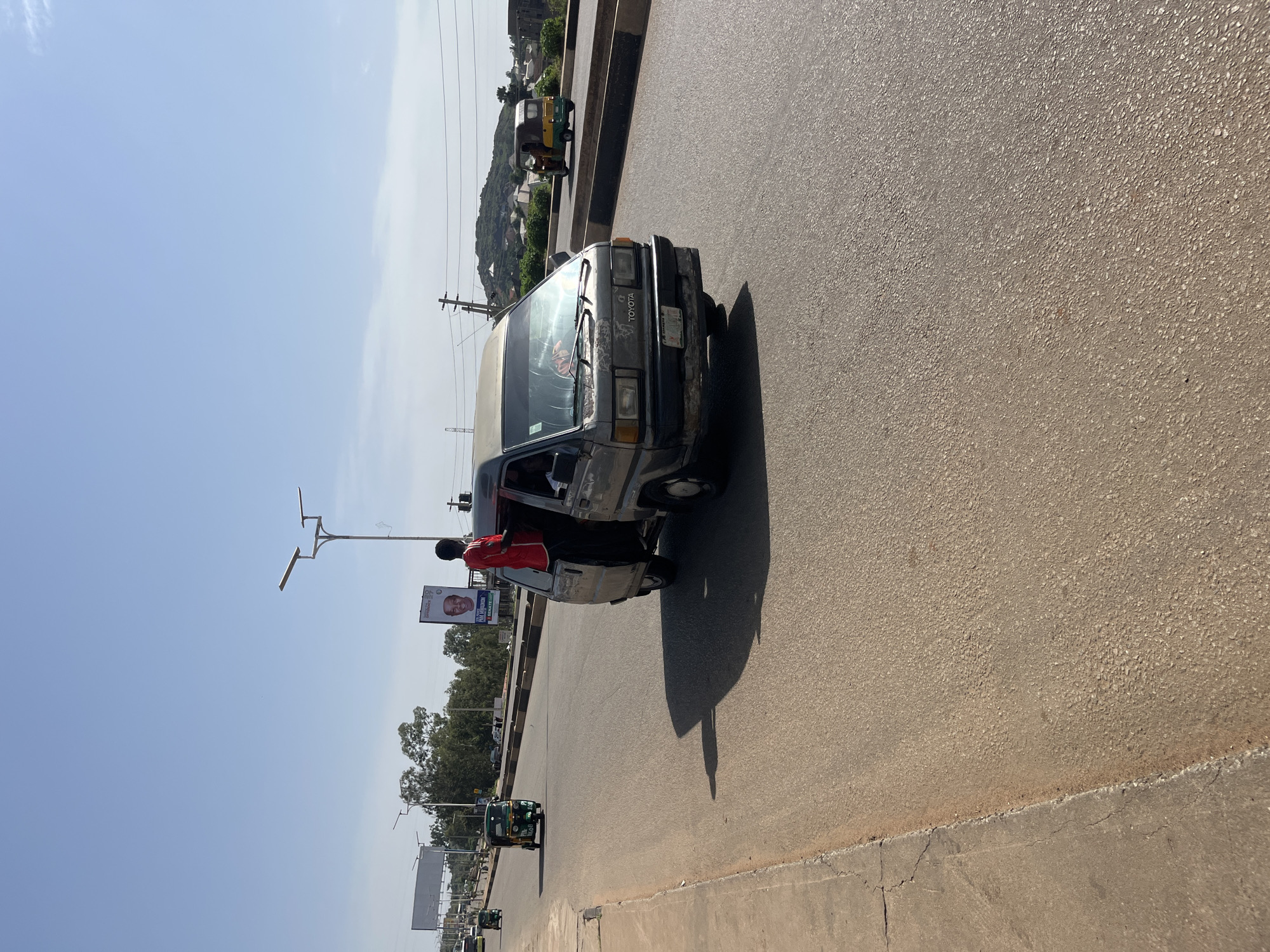 In the city of Gura Top, Nigeria, a bustling road is captured under a clear blue sky. A weathered gray van, possibly a Toyota, is the central focus, its side door open as a young man in a red shirt and black pants leans out. He appears to be either a passenger or an attendant, possibly engaged in signaling or attending to someone outside the vehicle.
The van is on a paved road, with a concrete sidewalk to its left. In the background, more vehicles are visible, including a bright green and yellow auto-rickshaw, a common mode of transport in Nigeria, moving away from the van. Further down the road, other vehicles and a billboard are discernible.
The environment suggests a warm, sunny day, with long shadows cast by the vehicles. The scene conveys a sense of everyday life and movement in a Nigerian urban setting.