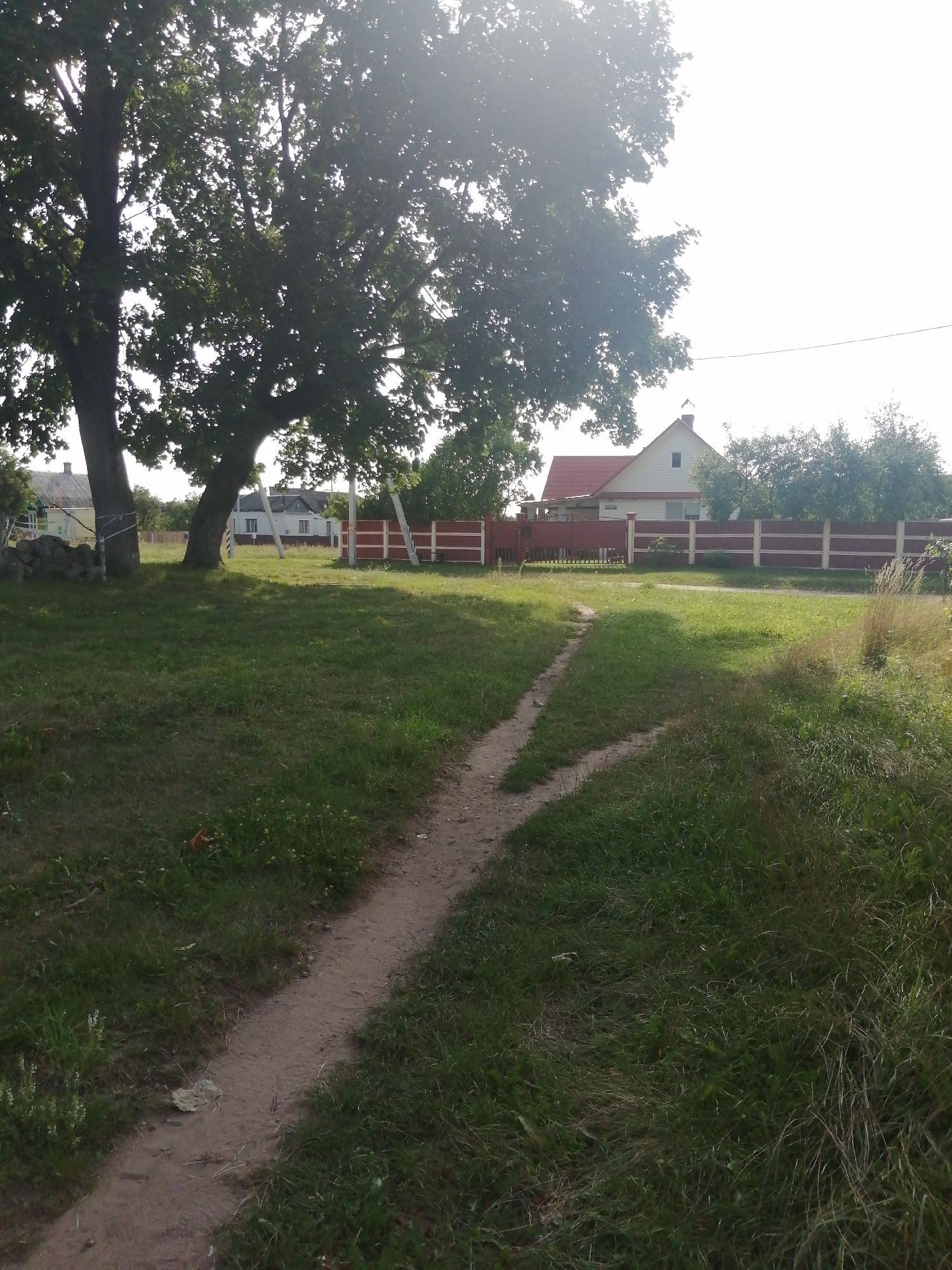 The image shows a path leading through a grassy area towards a red and white fence, with a house visible behind it. Large trees with dense foliage are present on the left side of the path. The sky is bright and slightly hazy, suggesting it may be daytime with the sun high. The overall scene depicts a rural or suburban setting. Without specific identifying landmarks or contextual information, it is not possible to definitively determine the exact location within Slonim, Belarus.