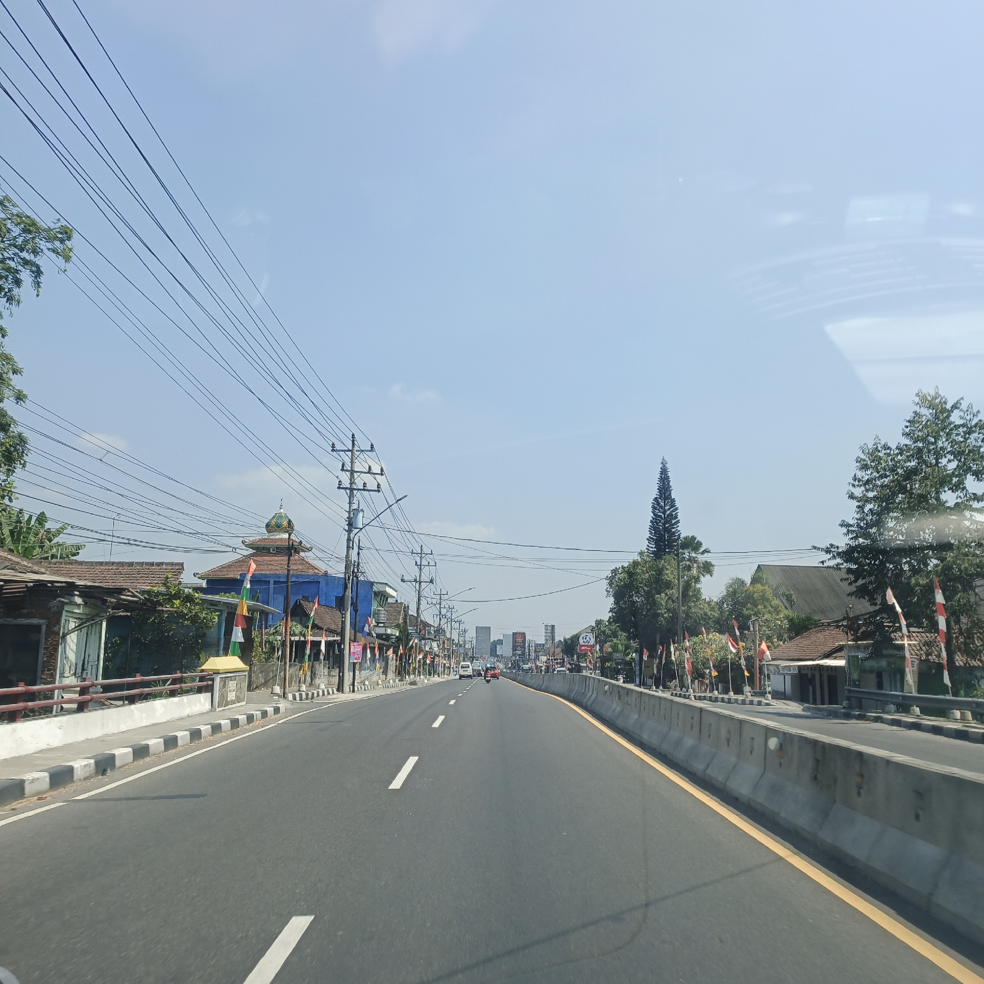 The image captures a view from a vehicle traveling along a paved road in Secang, Indonesia. The road is divided into lanes by white dashed lines and bordered by a concrete barrier with a yellow line. On the left side of the road, there are buildings with tiled roofs, some with visible brickwork, and utility poles with numerous electrical wires strung between them. A blue building with a dome-like structure and decorative flags is visible. The right side of the road also features buildings and trees, with more flags lining the barrier. Several vehicles, including cars and motorcycles, are seen on the road ahead. The sky is clear blue with a few wispy clouds. The overall scene depicts a typical urban or suburban street in Indonesia on a sunny day.