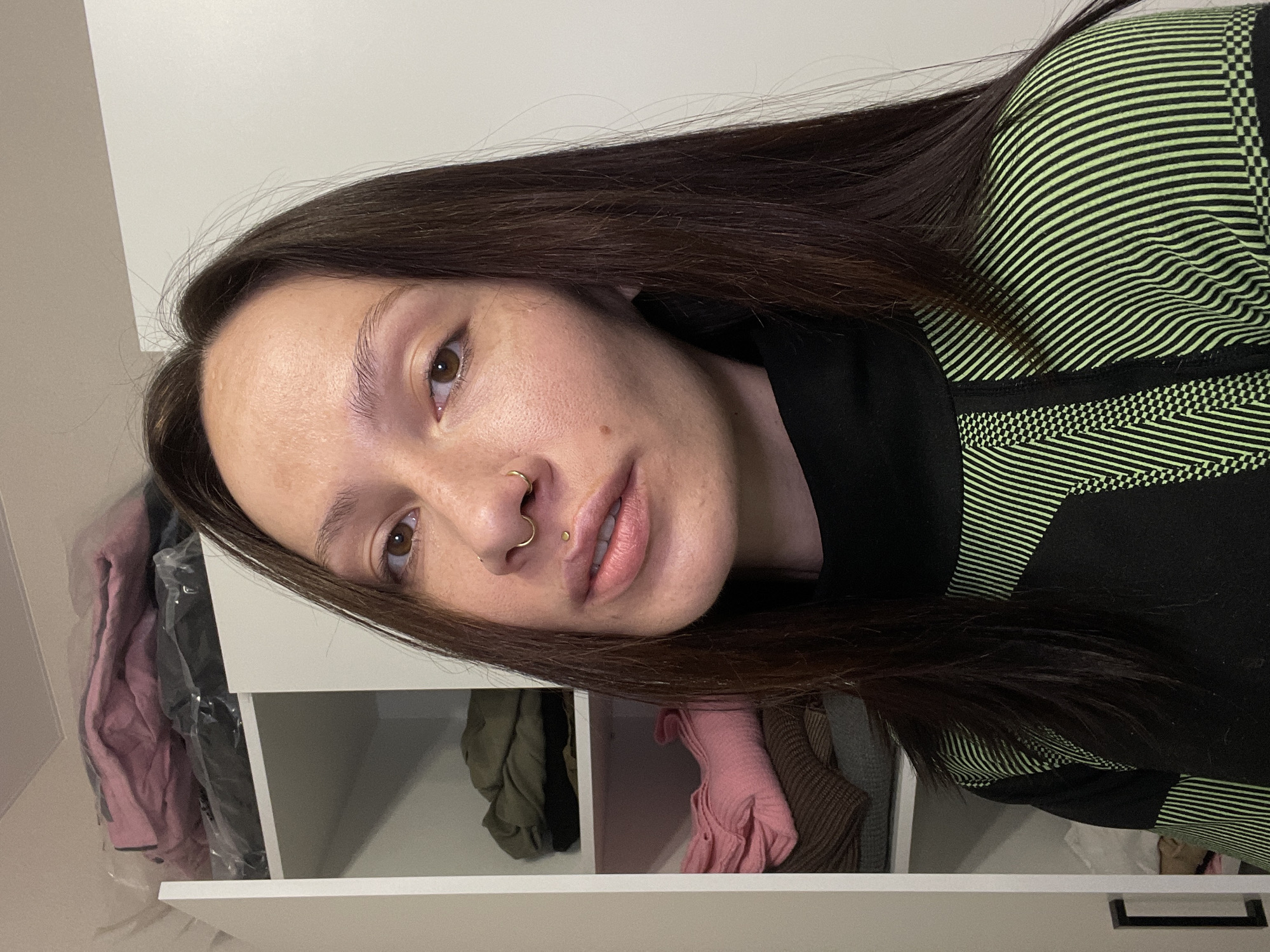 A close-up portrait captures a woman with long, dark brown hair and brown eyes, looking directly at the viewer. Her head is tilted, and she has fair skin, a gold-colored septum piercing, and a small gold stud in her philtrum. She wears a black long-sleeved top with distinctive neon green horizontal stripes and geometric patterns. The stripes vary in width and density, and some sections feature a small checkered pattern.
The background reveals light-colored shelving units or a closet, with various folded or hanging garments visible inside. These include items of clothing in shades of pink, dark green, and brown, some of which are encased in clear plastic. The setting is an indoor room, possibly a bedroom or dressing area, located in Pervomaysky, Russia. The lighting is soft and even, suggesting an indoor environment. No specific activity is taking place beyond posing for the portrait, and there is no visible text.