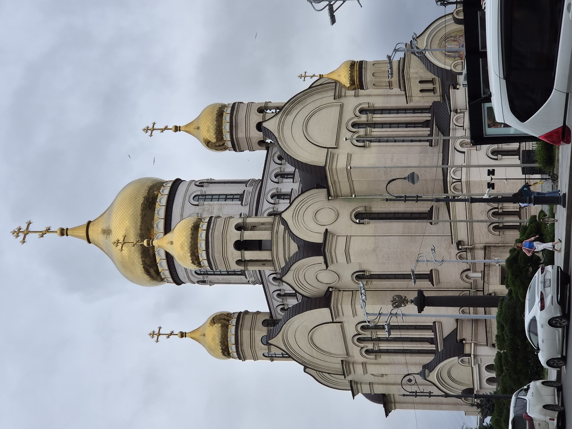This is a photograph of the Cathedral of the Intercession of the Theotokos in Vladivostok, Russia. The cathedral is a large, pale yellow building with multiple golden domes, topped with crosses. The sky is overcast and grey. In the foreground, there are several cars parked on the street, including a white minivan and a white sedan. A woman in a pink and white striped shirt and white pants is walking a dog on the sidewalk. Another person is standing on the sidewalk with a scooter. Trees and bushes are visible behind the sidewalk. Streetlights and a dark pillar with a double-headed eagle emblem are also present.