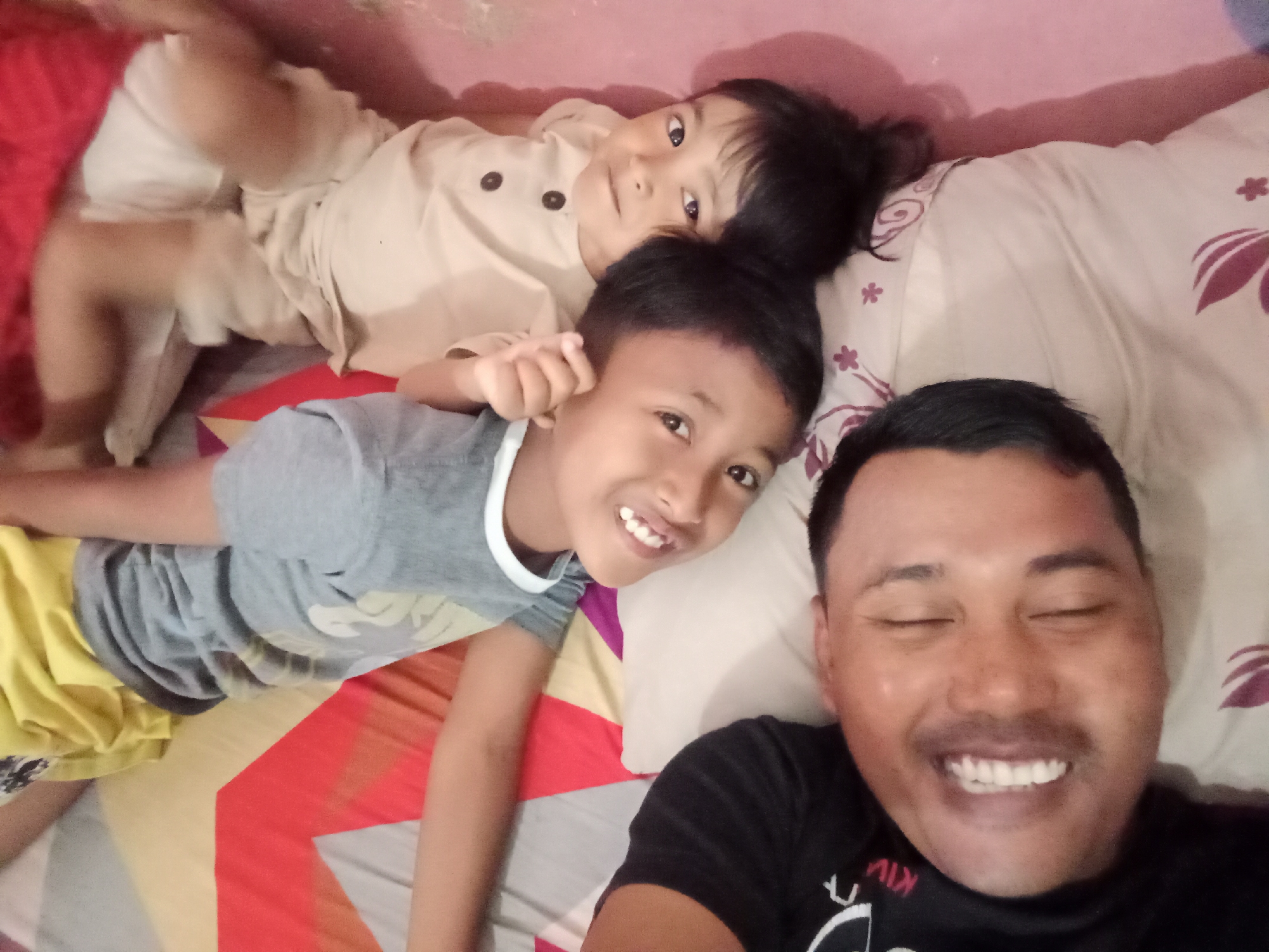 In the city of Bloro, Indonesia, three individuals are captured in a selfie from a high angle, lying on a bed. The person in the foreground, on the right, is an adult male with closed eyes and a wide smile, wearing a black t-shirt. To his left, a young boy with short dark hair, a grey t-shirt, and yellow shorts smiles at the camera. Behind him, a younger child with dark hair and a light-colored shirt is partially visible, also looking towards the camera. The bed is covered with a light-colored sheet featuring a floral pattern in shades of burgundy and white, and a pink wall serves as the background. The overall scene depicts a casual, familial moment.
