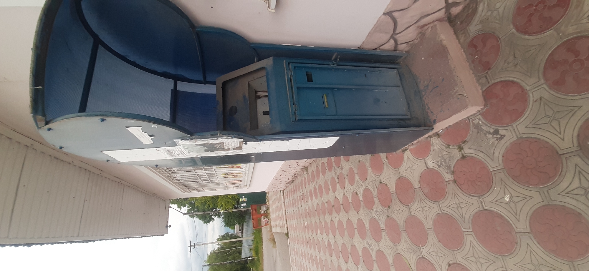 The image depicts an outdoor scene in Yessentukskaya, Russia, possibly near a building with a covered entryway. The main subject is a blue metal structure, possibly a shelter or enclosure, with a translucent blue roof. Inside this structure, a blue metal cabinet or safe is visible, appearing somewhat weathered and dusty. There are some papers or labels attached to the underside of the shelter's metal frame.
The ground in front of the structure is paved with decorative tiles featuring a repeating pattern of circular motifs in red and beige. To the left of the structure, a section of a building with a light-colored siding and a metal grate is visible. In the background, trees and utility poles suggest a residential or semi-urban area. The sky is overcast, indicating a cloudy or possibly rainy day.
There is no discernible activity or event taking place. The scene appears static and focuses on the stationary objects. No people are visible in the image. The overall impression is one of a functional outdoor space that might be associated with a building or a public utility. No clear text is legible within the image.