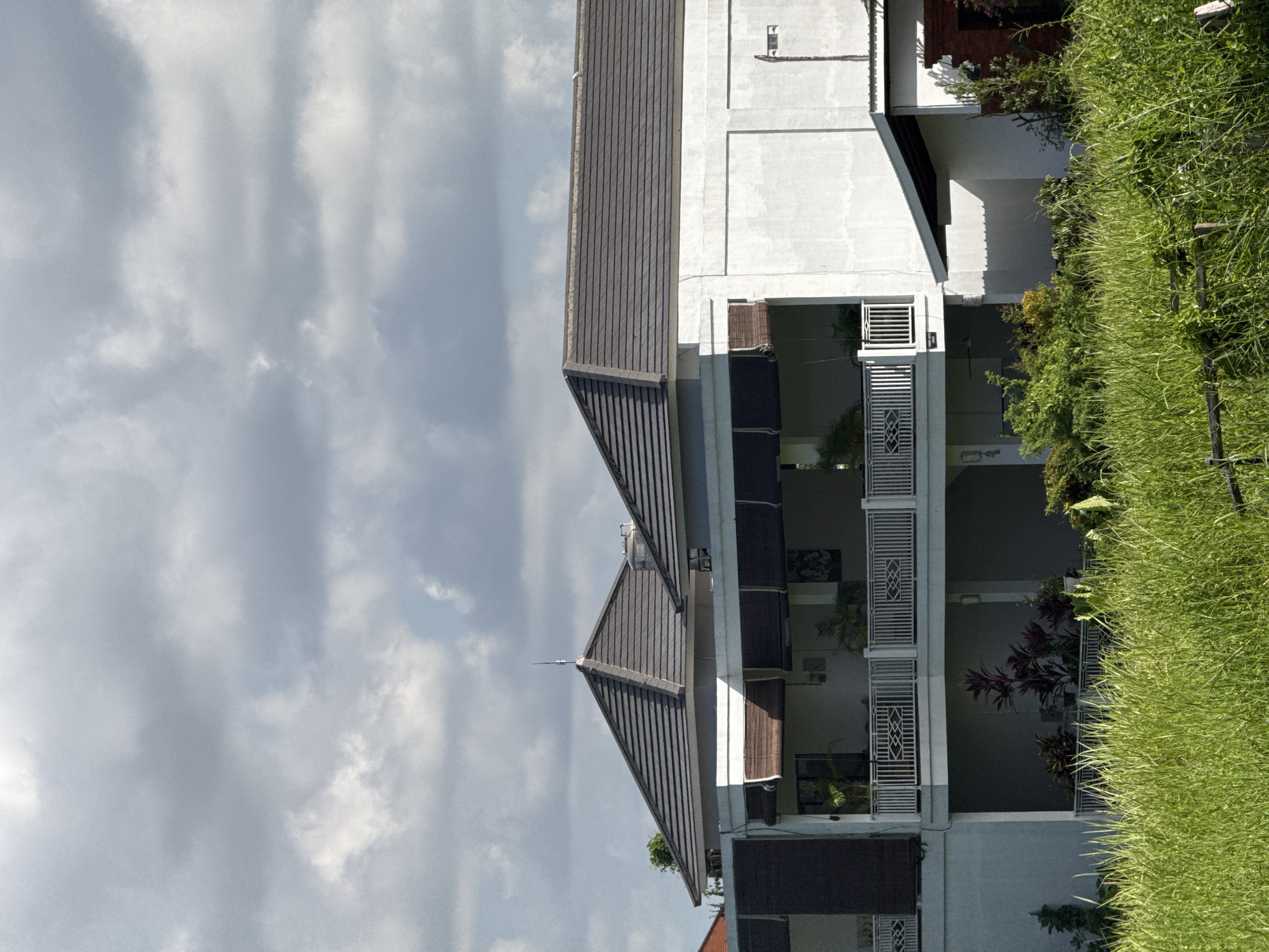 The image presents a view of a building, characterized by a grey tile roof and white exterior walls, under a cloudy sky. The building's design incorporates balconies with white railings. Lush green grass and plants grow near the structure, suggesting a residential or community setting. The scene appears to be located in North Kuta, Indonesia.