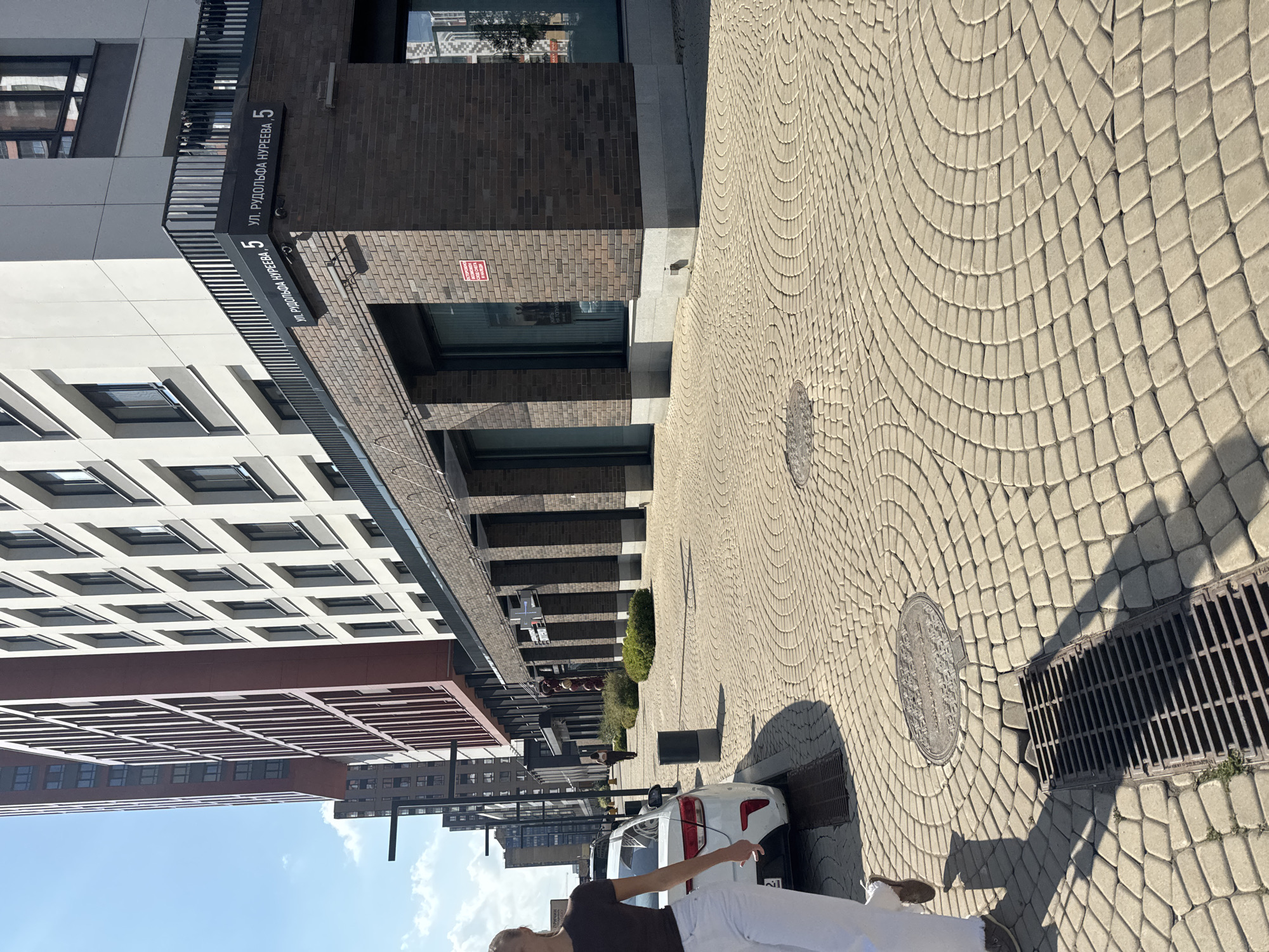The image depicts a street scene in Ufa, Russia, specifically on Rudolfa Nureyeva Street. The foreground is dominated by a patterned cobblestone sidewalk leading towards modern buildings. A white car is parked on the left side of the frame, and a person, partially visible from the side, is walking on the sidewalk. The buildings in the background are contemporary, with a mix of brick and white facades, featuring numerous windows and balconies. A street sign reading "ул. Рудольфа Нуреева, 5" is visible on one of the buildings, confirming the location. The environment appears to be a well-maintained urban area with clear blue skies overhead.