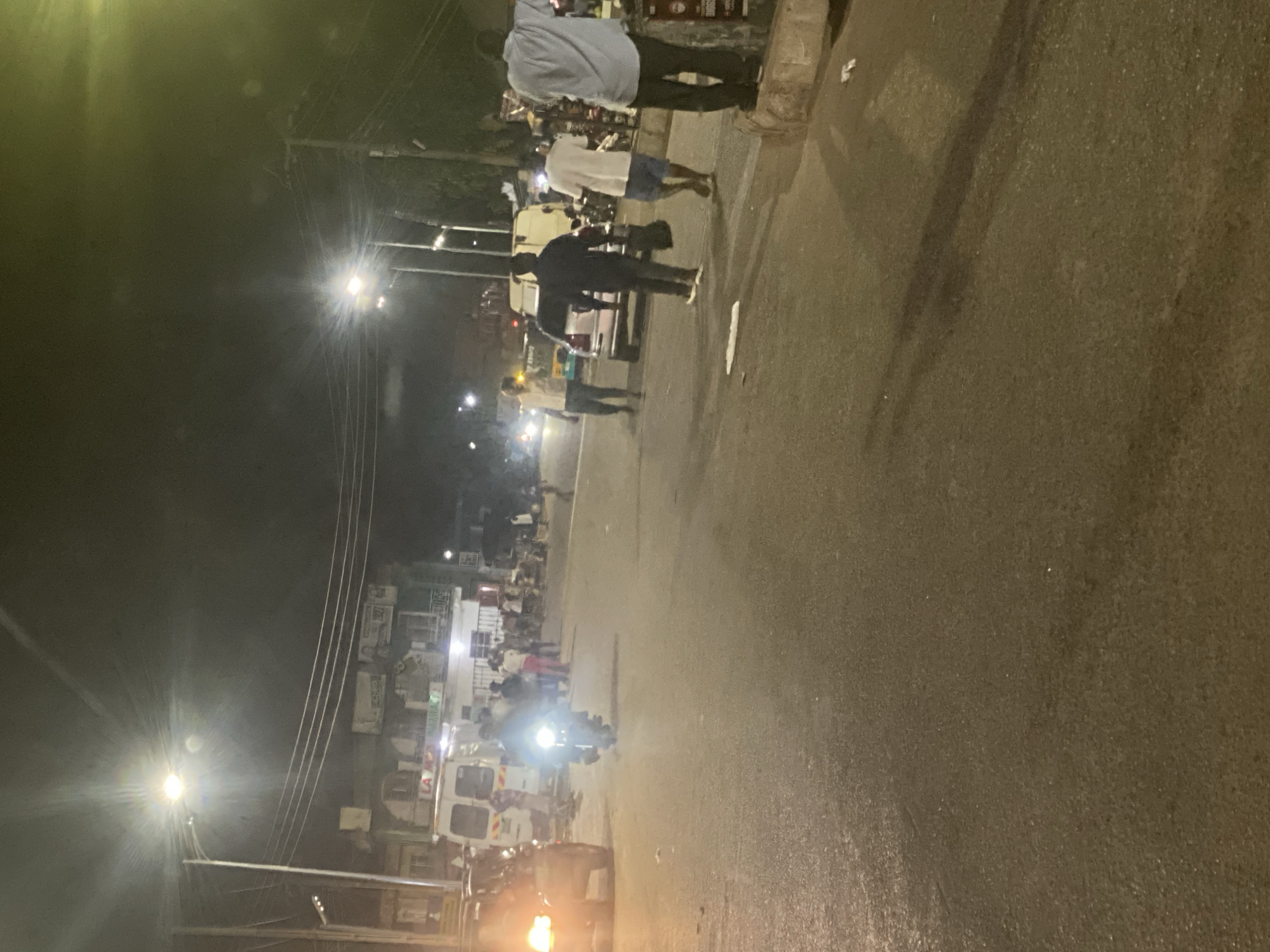 At night in Gwong, Nigeria, a street scene shows people walking on the sidewalk near an ambulance. The street, illuminated by streetlights and vehicle headlights, has an asphalt surface. The buildings in the background are lit up.