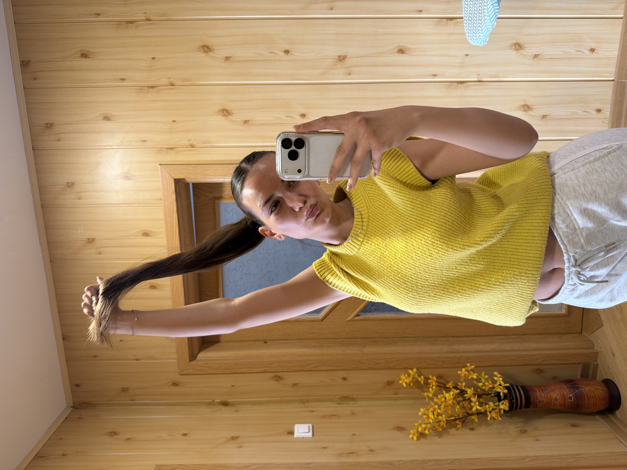 A woman with dark hair pulled back in a ponytail is taking a selfie in a room with wood-paneled walls. She is wearing a yellow sweater vest and light-colored sweatpants. She is holding a white smartphone with multiple camera lenses up in her right hand, with a slight pout on her face. Her left arm is extended, holding the end of her ponytail. The room has a door with a wooden frame and a light switch on the wall near the floor. There is a brown vase with yellow flowers on the floor. The location appears to be indoors, possibly a home. There is no immediate evidence to confirm the location is Buhovo, Bulgaria.
