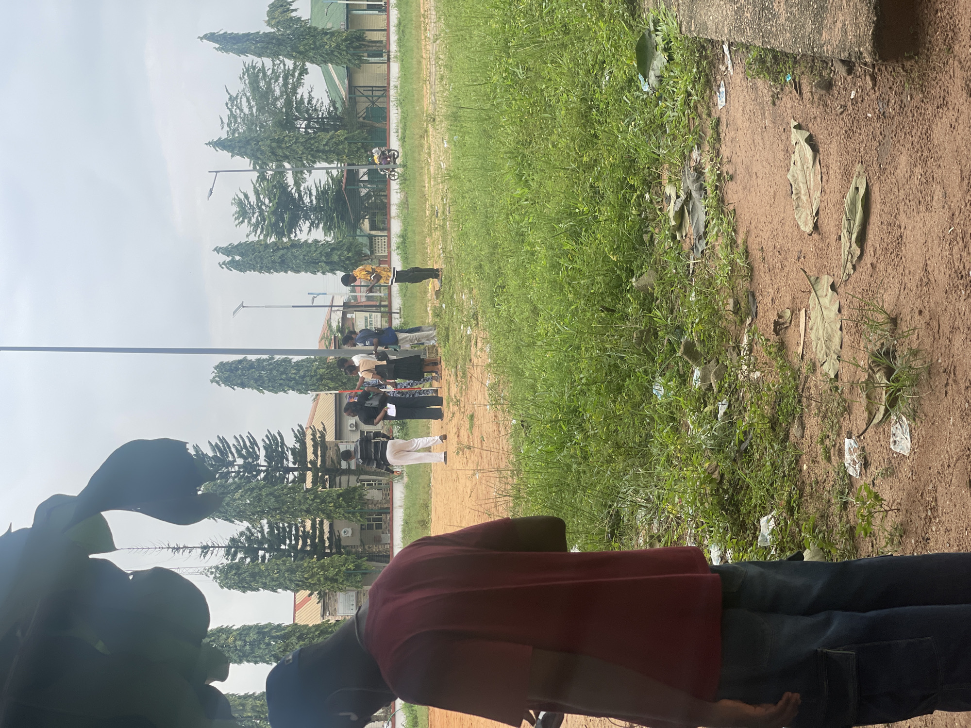 In Ihiagwa, Nigeria, on an overcast day, a group of people are gathered outdoors. In the foreground, a person wearing a maroon t-shirt and blue jeans is seen from behind, facing away from the camera. Further back, several other individuals stand on a dirt and grass area. Behind them is a building with an off-white facade and a green roof, flanked by tall, slender trees. A motorbike with a rider is visible in the middle ground, on a road adjacent to the grassy area. The environment appears somewhat unkempt, with scattered debris and dried leaves on the ground. The overall scene suggests a public space with a casual gathering of people.