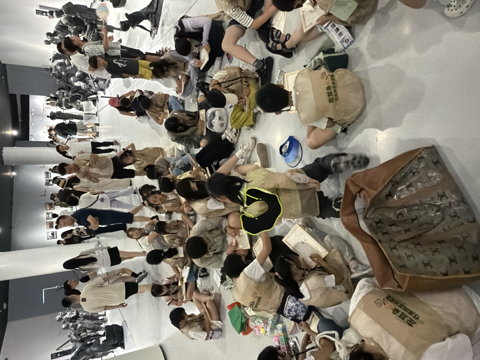 This image depicts an indoor scene within an art gallery or museum, identified as being in the Chongqing High-tech Zone, China. The space features a light grey floor, white walls, and a dark ceiling equipped with bright overhead and track lighting. Large white cylindrical columns are visible in the mid-ground.
Approximately 20-30 children, primarily between 6 and 12 years old, are seated on the floor in scattered groups across the foreground and mid-ground. Many are wearing tan vests with the Chinese characters "左耳朵 行动艺术营" (Left Ear Action Art Camp) printed on the back. These children are actively engaged in drawing or writing on small pads or canvases, using pens or pencils. Various art supplies, including colored items in plastic bags, a blue visor, and small personal bags, are arranged around them on the floor. One child in the mid-ground is wearing a distinctive black and yellow U-shaped accessory around their neck.
Multiple adults are present. Some adults are seated on the floor among the children, appearing to supervise or participate. In the background, numerous other adults are standing and walking, observing the art exhibits displayed throughout the gallery. Their attire ranges from casual to semi-casual.
The art exhibits include multiple dark-colored sculptures depicting human figures in various poses, some grouped to form scenes. These are visible on the left side and in the far background on the right. Framed artworks are displayed along the walls in the background. A large brown tote bag with a repeating "CC" logo pattern is prominently positioned on the floor in the immediate foreground.