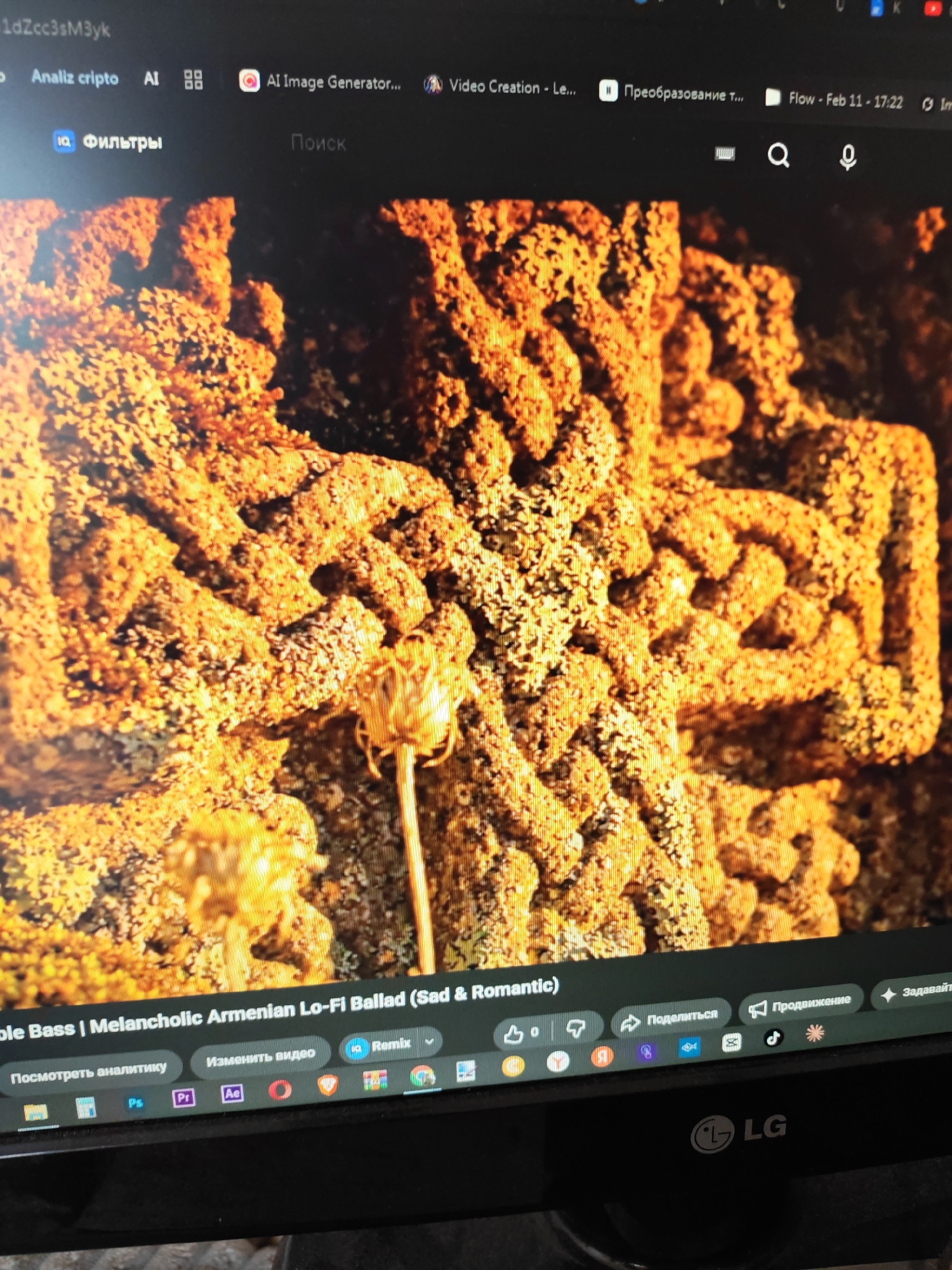 The image shows a close-up of a computer screen displaying a YouTube video or similar media player. The video content features a textured, relief-like carving in a warm, golden-brown hue, possibly stone or wood, resembling an intricate, stylized cross with floral or leaf motifs. The carving is highly detailed and appears weathered or ancient. In the foreground of the video, a dried flower or seed pod on a slender stem is visible.
The computer's interface is partially visible, with a dark taskbar at the top containing text like "Analiz cripto AI", "AI Image Generator...", and "Flow - Feb 11 - 17:22". Below the video, the bottom taskbar shows icons for media playback controls, a video title "ble Bass | Melancholic Armenian Lo-Fi Ballad (Sad & Romantic)", and options like "Посмотреть аналитику", "Изменить видео", "Remix", "Поделиться", "Продвижение", and "Задавайте". There are also icons representing various applications such as Adobe Photoshop, Adobe Premiere, and Adobe After Effects. The bottom edge of the monitor displays the "LG" logo.
The user-provided information about the location being Vanadzor, Armenia, is external to the visual content of the image itself. The image itself does not contain any visual cues to identify its geographical location or any specific details about Vanadzor.