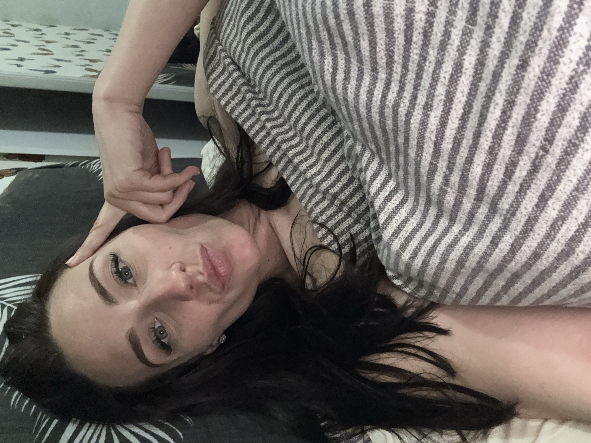 A woman with dark hair is lying on a patterned surface under a striped blanket. She is looking at the camera with her head tilted. Her hand is resting on her forehead. The location cannot be determined from the image.