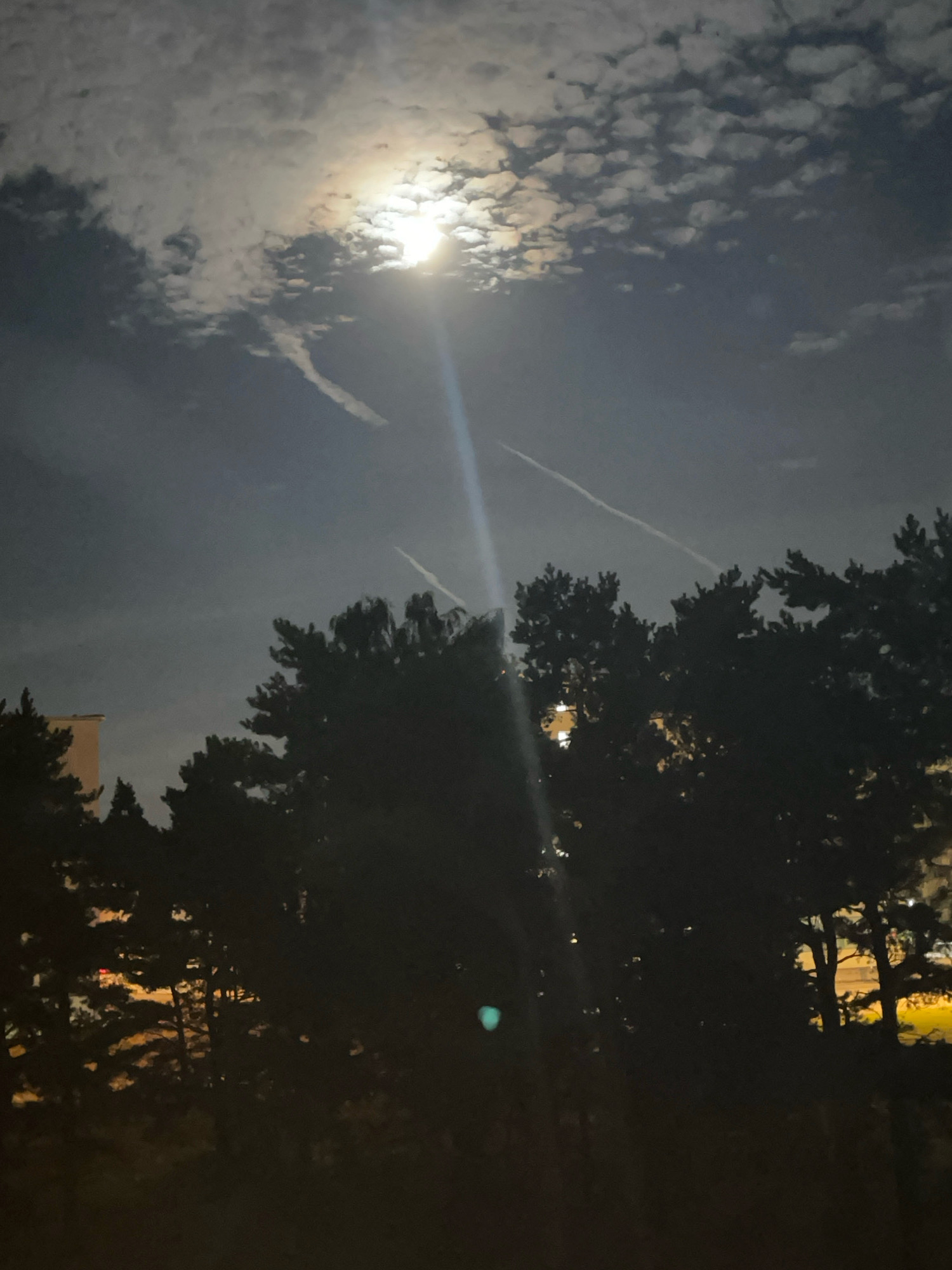 The image depicts a nighttime scene, with the full moon partially obscured by clouds, casting a bright light that creates a visible ray. Several contrails, likely from airplanes, stretch across the sky. Silhouetted against the moonlit sky are the tops of pine trees. To the left, a portion of a building is visible. The foreground shows more trees in silhouette. The overall atmosphere is dark, with hints of artificial light from what appears to be a city in the distance. There are no people or visible animals in the image. The scene is not identifiable as being specifically in Tallinn, Estonia, or any other particular city.