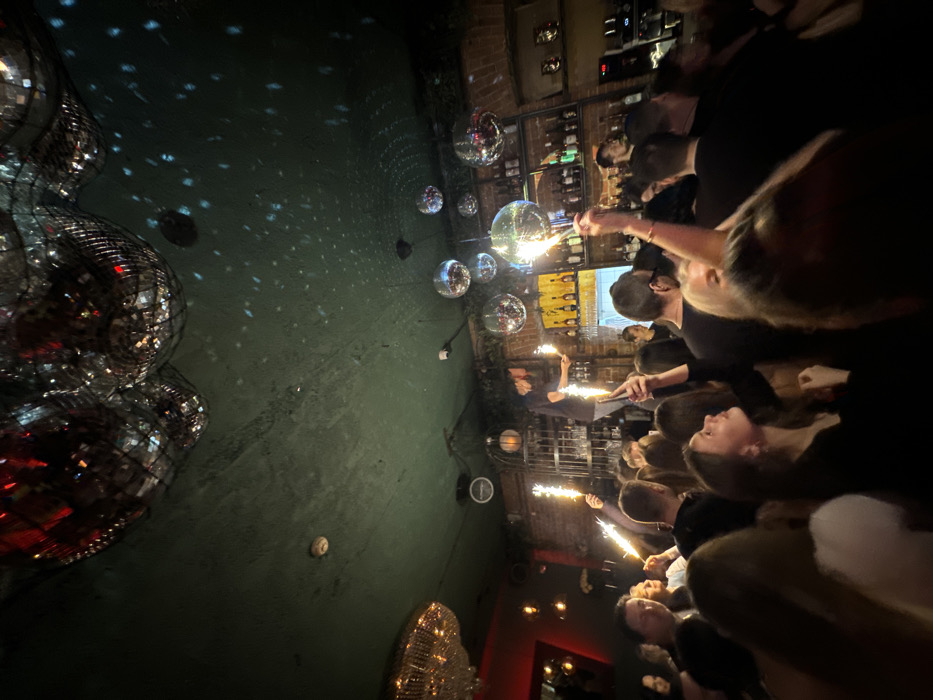 The image depicts the interior of a crowded, dimly lit establishment identified as Butler the Bar, located in Moscow, Russia. The ceiling, painted a dark green, features multiple suspended disco balls of various sizes, reflecting light to cast scattered blue and white patterns. A large, ornate chandelier hangs on the left. A circular smoke detector is affixed to the ceiling.
In the background, exposed brick walls are visible behind a well-stocked bar, lined with numerous bottles of alcohol. A male individual, appearing to be staff, stands atop a metal cage-like structure behind the bar, holding two actively lit handheld sparklers.
The foreground and mid-ground are densely populated with numerous individuals, predominantly young adults. Multiple members of the crowd are holding actively burning handheld sparklers, producing bright, upward-streaming sparks. Many individuals are observed looking upward, towards the sparklers and ceiling. At least two individuals in the mid-ground are holding smartphones aloft, seemingly recording the scene. The overall lighting is dim, supplemented by the bright light from the sparklers and reflections from the disco balls.