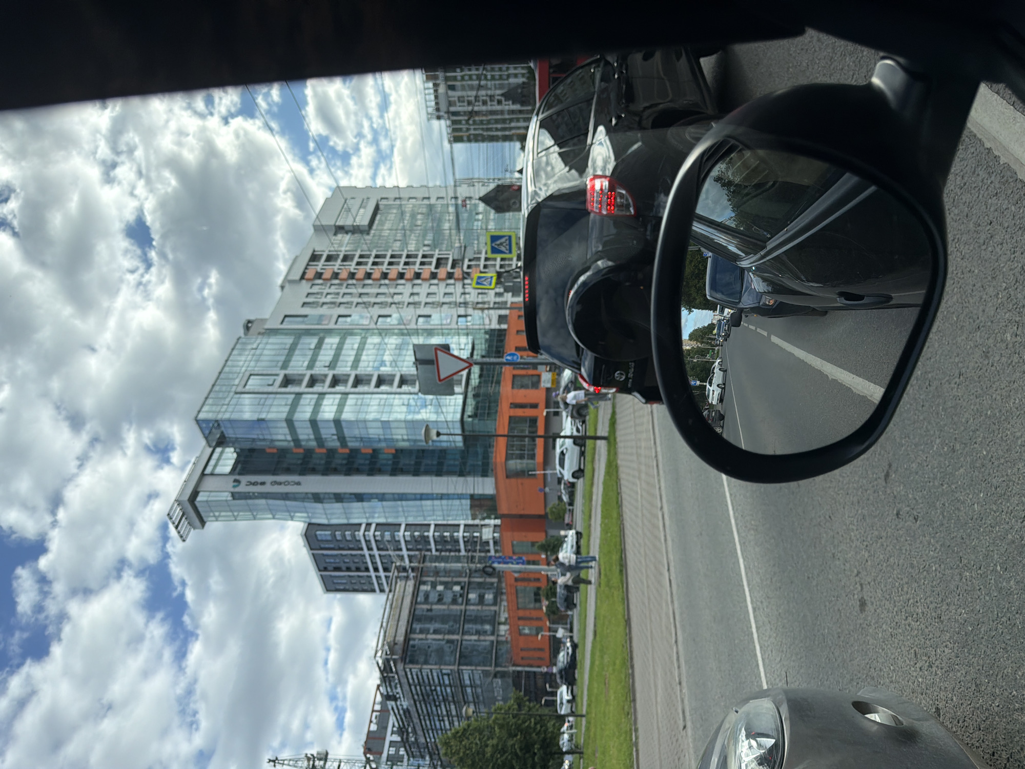 The image shows a view from inside a vehicle, looking forward and to the side mirror. In the foreground, a portion of the vehicle's dashboard and side mirror are visible. The side mirror reflects a line of cars driving on a city street, with a dark SUV in the immediate reflection. Beyond the reflection, the street is populated with several other vehicles.
In the background, modern residential and office buildings rise against a sky filled with white, fluffy clouds. These buildings have a contemporary architectural style, featuring glass facades and varying colors, including orange accents on some structures. A construction crane is also visible in the distance on the left side of the frame.
The street in front of the buildings appears to be a multi-lane road with pedestrians walking on a sidewalk to the left. Traffic signs and streetlights are present. The overall environment suggests an urban setting on a cloudy day. Based on the architectural style and urban landscape, this scene is likely located in Izhevsk, Russia, possibly in a newly developed area of the city.
