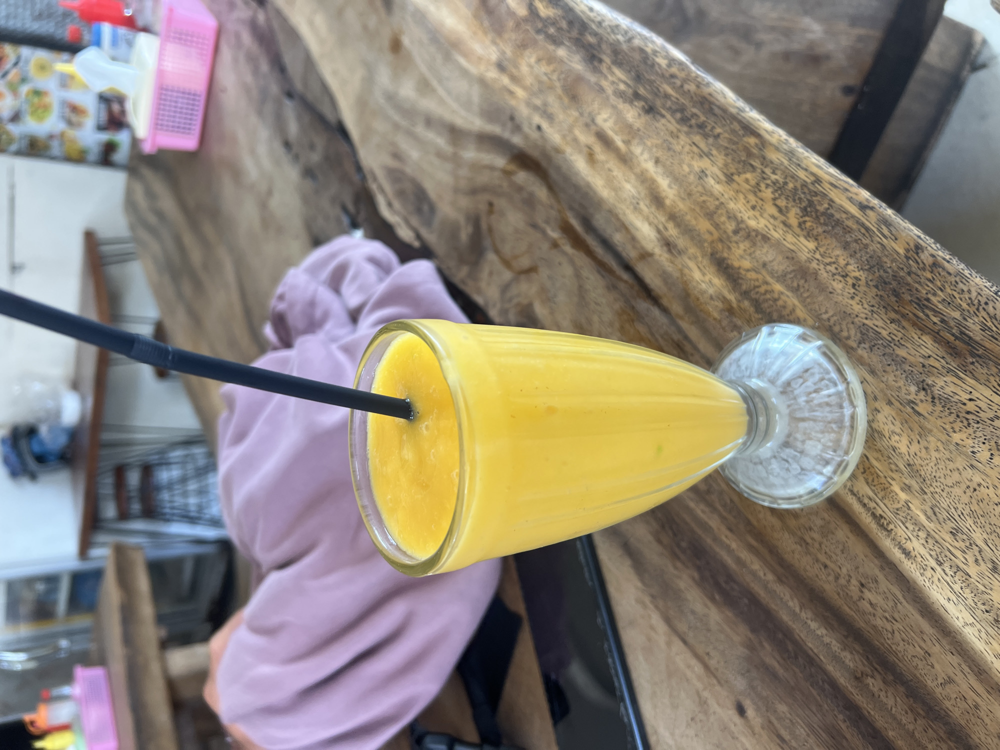 A tall glass filled with a yellow, frothy beverage, likely a mango smoothie, sits on a rustic wooden table. A black straw protrudes from the drink. The table's surface is textured and shows signs of wear. In the background, a person in a lavender-colored garment is partially visible, seated. Further back, other tables and chairs are present, suggesting an outdoor or casual indoor dining setting. A pink plastic basket containing items is visible on a table in the mid-ground. The overall impression is a casual moment captured at a cafe or restaurant in Kuta, Indonesia, perhaps a beachside establishment given the wooden furniture and relaxed atmosphere.