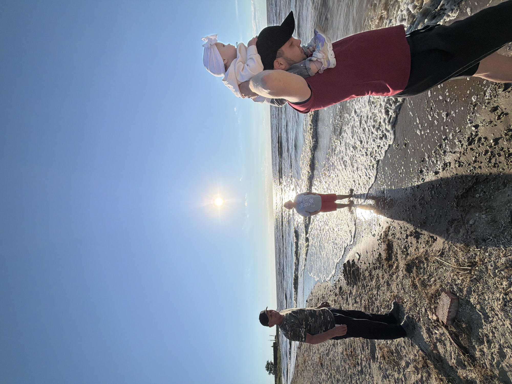 In the city of Port-Katon, Russia, on a beach bathed in the golden light of a setting sun, three individuals are captured in a moment of shared joy. The sky above is a canvas of clear blue, transitioning to a softer hue as it meets the horizon. The sun, a radiant orb, casts a shimmering path across the gentle waves that lap at the shore.
In the foreground, a man stands with his back to the camera, a black baseball cap shielding his eyes. Perched on his shoulders is a baby, wrapped in a white bonnet and a light-colored outfit, looking out towards the vast expanse of water. The man's attire consists of a deep red t-shirt and dark shorts.
To the left, another man stands facing the sea, his camouflage t-shirt a stark contrast to the sandy terrain. He wears a dark baseball cap and black pants. Further back, a third figure is visible, standing by the water's edge, their lower half obscured by the shimmering reflections of the sun. The beach itself is a mixture of sand and small pebbles, with scattered bits of dried seaweed and debris. A single red brick lies prominently on the sand, a solitary object amidst the natural elements. The scene evokes a sense of peace and familial connection, a quiet interlude by the water.