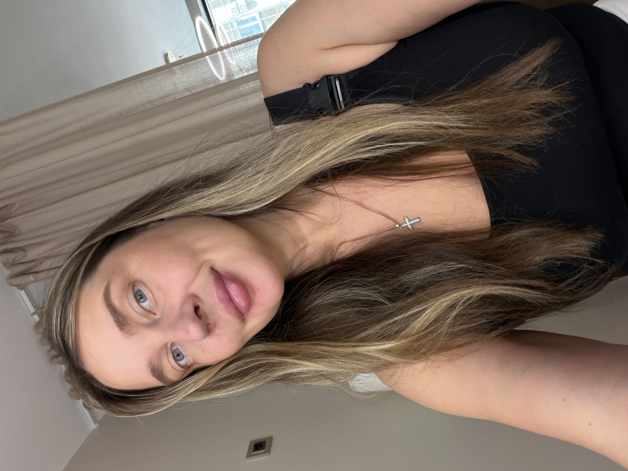 The image shows a young woman with long, blonde-brown hair and blue eyes. She is wearing a black tank top and a gold necklace with a cross pendant. She has a nose ring. The background is a room with beige curtains and a window with a view of a city skyline. There is a light fixture visible in the background. The woman is looking at the camera and smiling slightly. The image appears to be a selfie taken indoors. There is no indication of the location being Moscow, Russia.