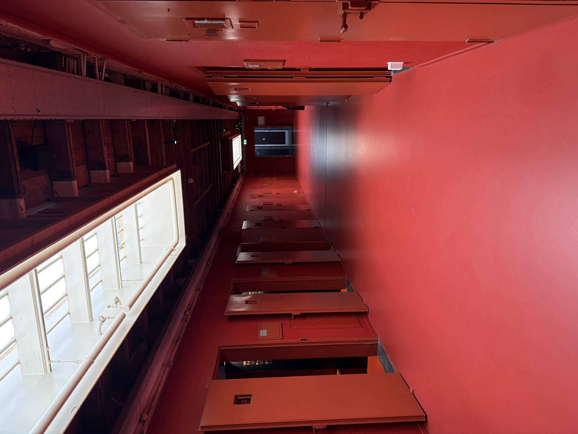 This image shows a long, narrow red hallway with numerous red doors on both sides. The doors appear to be cell doors, with small square windows on each. The floor is also bright red and appears to be a linoleum or similar smooth material. There are skylights in the ceiling, allowing natural light to enter the hallway, and fluorescent lighting fixtures are also visible. The hallway extends into the distance, where it appears to turn. This hallway is located in the former Bijlmer Prison in The Hague, Netherlands, which has been repurposed as a museum called 'Museum De Gevangenpoort'.