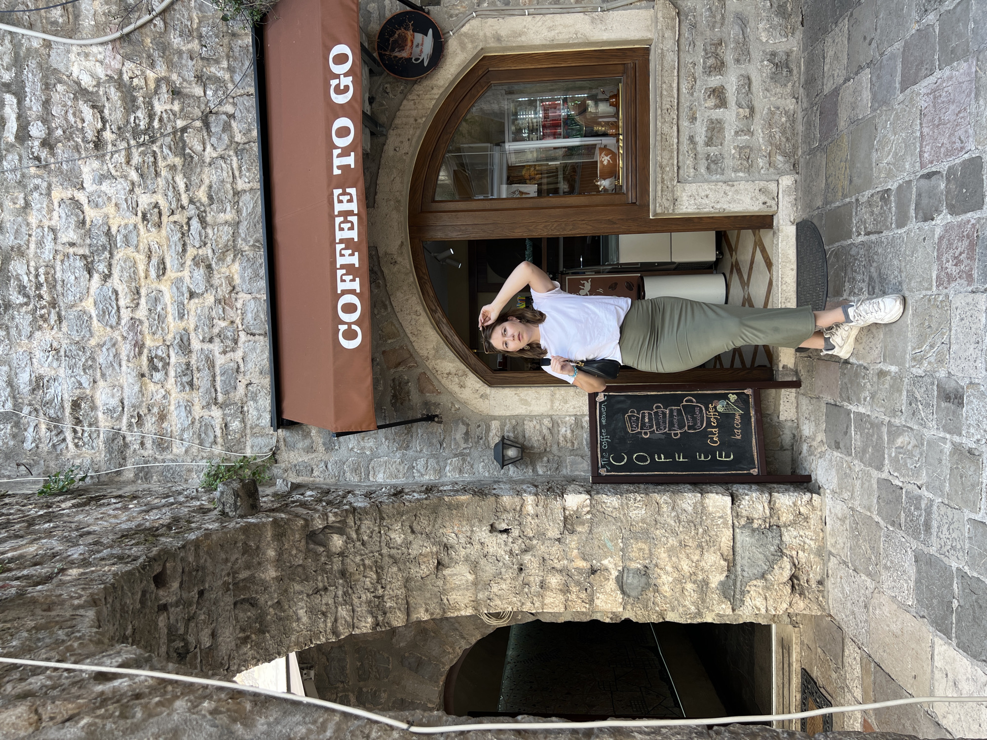 A young woman stands posing in a narrow, historic stone alleyway in Budva, Montenegro, next to the entrance of a "COFFEE TO GO" shop. The scene is bathed in bright daylight, suggesting a clear day.
The woman, with medium-length brown hair, wears a casual white t-shirt, a long olive green skirt, and white sneakers. She carries a black shoulder bag and holds sunglasses in one hand, looking directly towards the camera with a relaxed expression.
The coffee shop features a rustic brown awning displaying "COFFEE TO GO" in white capital letters. Above the awning, a round black sign shows a coffee cup image and the partially visible text "The coffee Heaven." The shop's entrance consists of an arched wooden-framed door with glass panels, through which parts of the interior, including drinks in a cooler, are faintly visible. To the right of the entrance, a black chalkboard sign lists various coffee offerings and treats.
The surroundings are characterized by rough-hewn, light-grey stone walls, forming part of ancient-looking structures. An archway made of similar stone is visible beneath the coffee shop, leading to another darker passage. A small black lamp is affixed to the stone wall, and some electrical wires run along the surfaces.
Visible text in the image includes:
- On the awning: "COFFEE TO GO"
- On the round sign: "The coffee Heaven"
- On the chalkboard: "COFFEE", "LATTE", "Espresso", "Cappuccino", "AMERICANO", "Hot CHOCOLATE", "Cold coffee", "Ice cream"