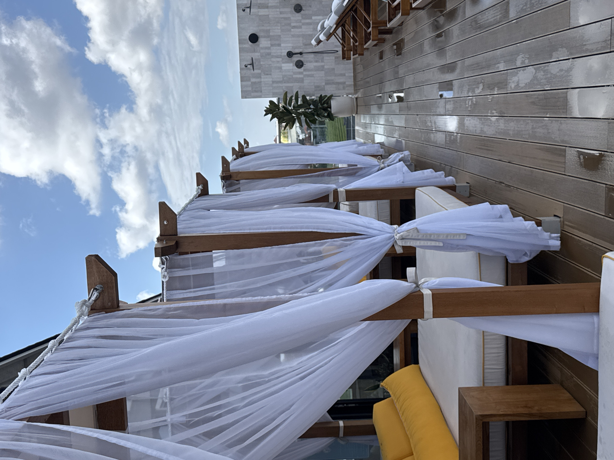 The image depicts a row of outdoor cabanas with white sheer curtains on a wooden deck in North Kuta, Indonesia. The cabanas feature wooden frames and are furnished with white mattresses and bright yellow cushions. The wooden deck appears wet, suggesting recent rain or cleaning. In the background, there are lounge chairs, outdoor showers with white tiled walls, and a potted plant. The sky is partly cloudy with blue patches. The overall scene suggests a resort or hotel setting, possibly a pool or beach area.