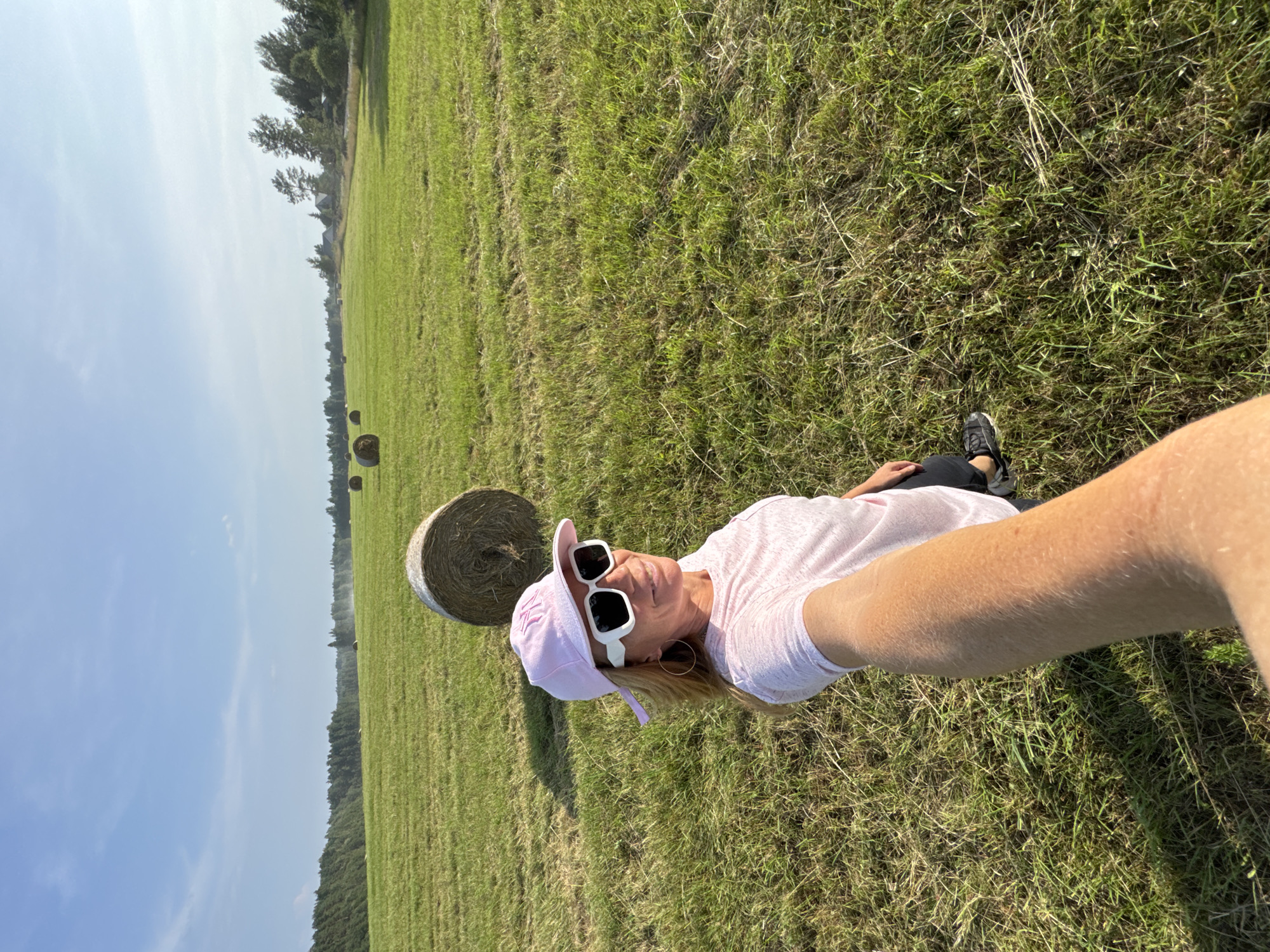 A woman is lying on her side in a grassy field with hay bales visible in the background. She is wearing a pink t-shirt, a light pink baseball cap, and white sunglasses. Her left leg is bent, and her right arm is extended, holding the camera. She is smiling at the camera. The scene appears to be a rural setting with a treeline in the distance. The sky is bright blue with some clouds, suggesting it is daytime and the weather is fair. The orientation of the image is portrait. There is no visible text in the image.