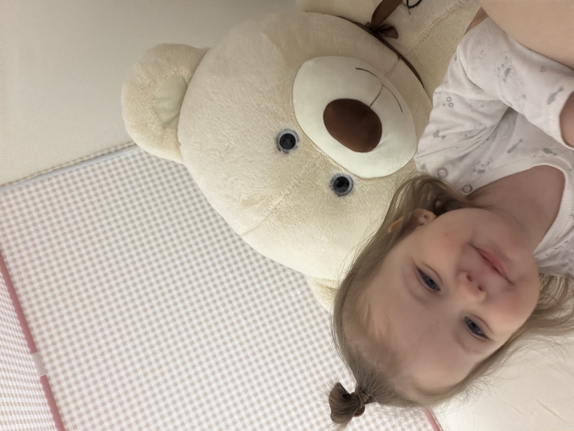 A young child with blonde hair tied in a small ponytail is lying on a white and beige checkered surface next to a large, cream-colored teddy bear. The child is wearing a white patterned shirt. The background includes a crib with similar checkered padding. The location is unknown and cannot be determined to be Reshetnikovo, Russia based on the image.