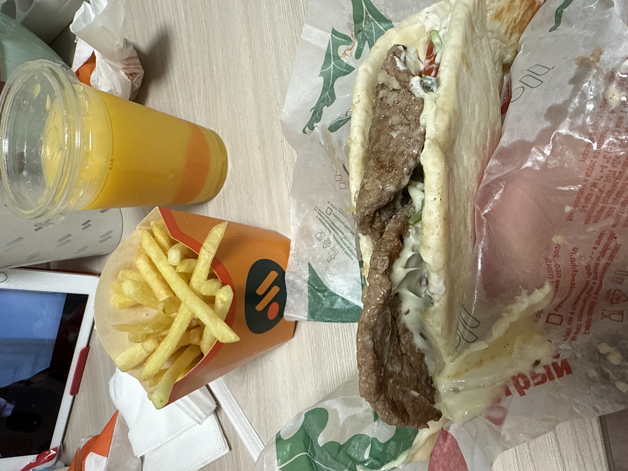 A close-up, overhead shot shows a meal on a wooden surface. In the foreground, a large sandwich is wrapped in paper, with a portion of the filling visible, including meat and creamy sauce. To the left and slightly behind the sandwich, a red and orange carton of french fries stands upright. To the right of the fries, a clear plastic cup filled with orange juice is visible. In the background, a tablet device is partially visible on the left side of the frame, and part of a white paper cup with a pattern is visible on the right. The lighting is bright, casting shadows on the wooden surface.