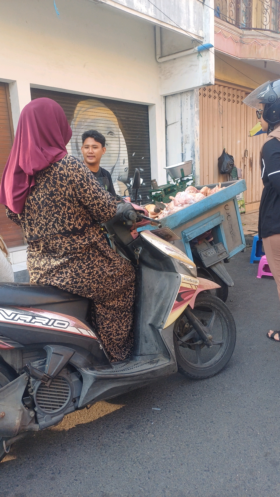 A woman wearing a maroon hijab and a black and gold patterned dress is seated on a dark-colored scooter, facing away from the camera. She is engaged in conversation with a young man standing beside her, who is wearing a black t-shirt. The scooter is parked on an asphalt road. Attached to the back of the scooter is a blue wooden crate overflowing with raw chickens. A green weighing scale is visible behind the crate. In the background, a building with a rolled-up metal shutter is visible. Further to the right, a person wearing a black jacket and helmet, and tan pants is standing. There are two colorful plastic stools on the ground to the right of the frame. The scene appears to be a street-side market or roadside stall. The overall lighting suggests it is daytime. The license plate of the scooter is partially visible and reads "L 2580 UG". The location is identified as Pekelingan, Indonesia.