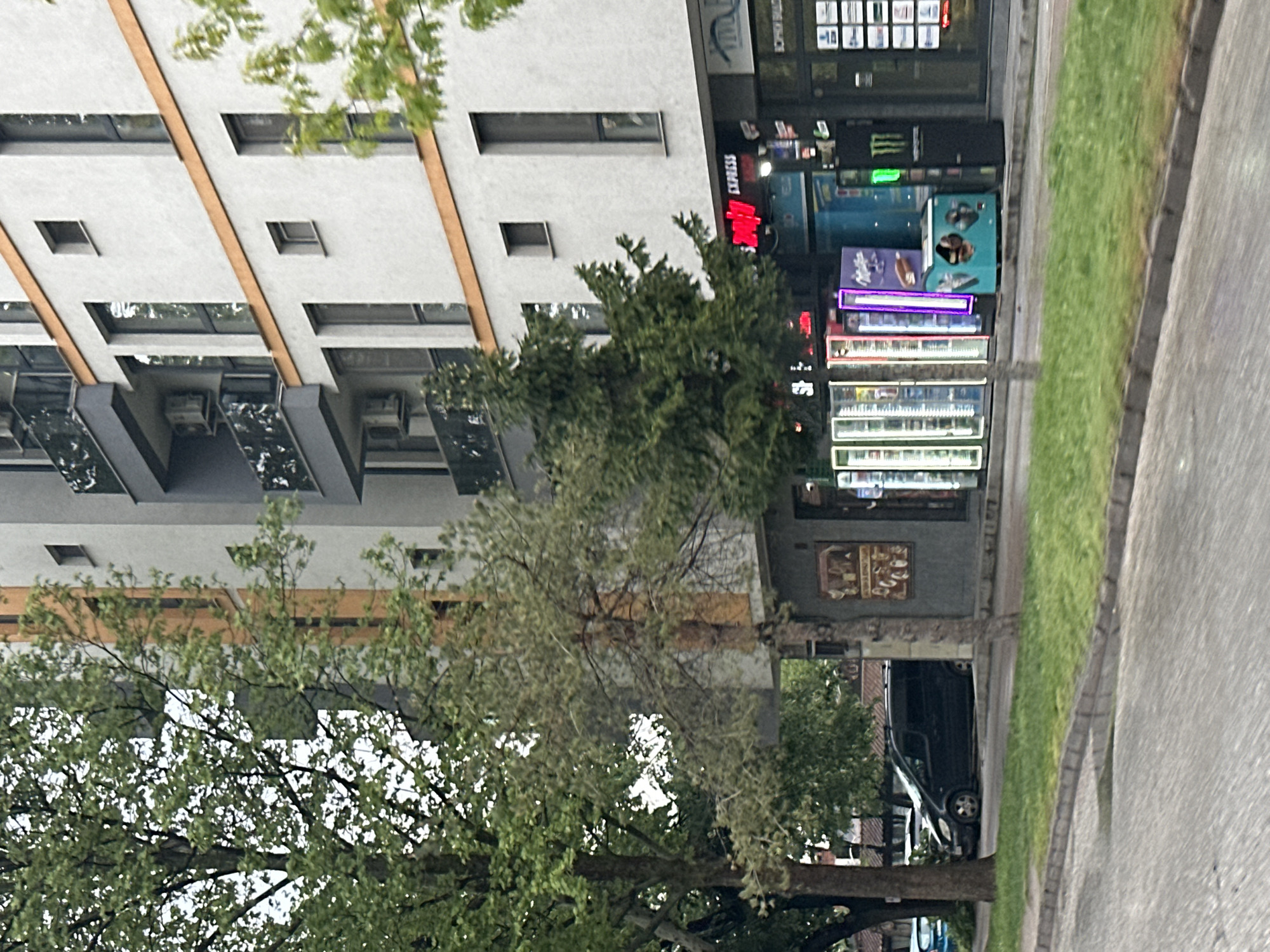 The image shows a street scene in Plovdiv, Bulgaria. A multi-story building is visible on the left, with modern architectural features. Adjacent to the building is a store or shop with "EXPRESS" displayed above the entrance. Vending machines or refrigerated displays with various products are in front of the store. Trees are visible in the foreground, partially obscuring the building and shop. A car is parked near the store. A sidewalk and a section of the road are visible on the right side of the image. The scene is set during daylight hours.