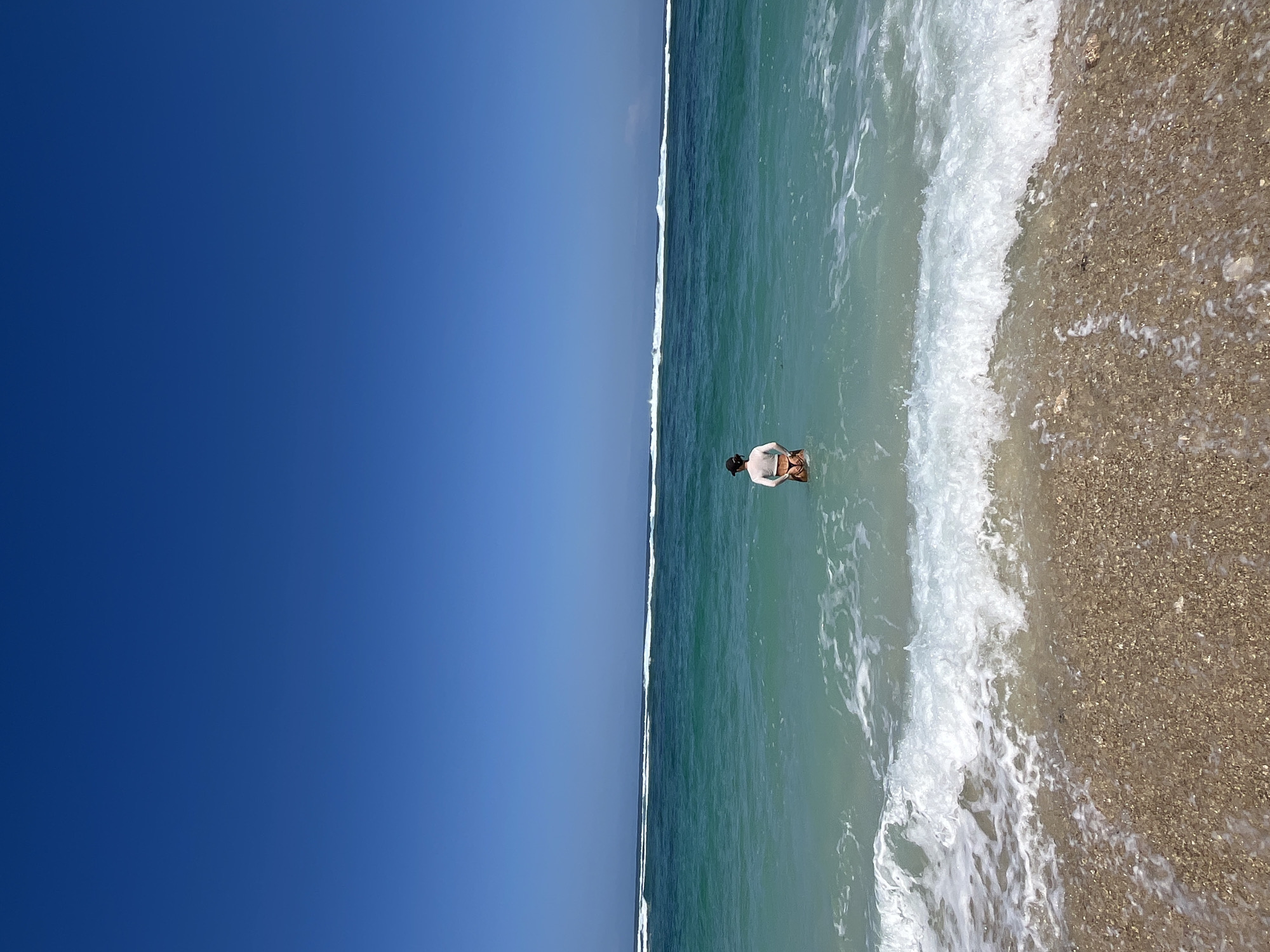 A person wearing a light-colored top and dark-colored bikini bottoms stands in the ocean in front of a beach in Kuta Selatan, Indonesia. The person is facing away from the camera, looking out at the sea. The water is a clear turquoise color with white waves crashing on the shore. The beach is covered in small, light-colored pebbles. The sky is a clear, bright blue with no clouds. The scene is tranquil and serene.