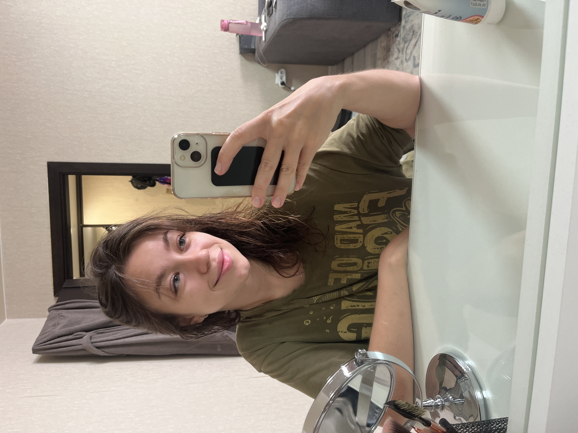 A young woman with medium-length brown hair and blue eyes is taking a mirror selfie. She is wearing a green t-shirt with text and graphics on it. She is sitting at a white vanity table with a mirror in front of her. To her left, a makeup brush is visible. In the background, a closet door is open, revealing a grey robe hanging on the door. Further in the background, a grey sofa and a pink water bottle are visible. The scene appears to be in a bedroom or dressing area. This image was taken in Chaikovsky, Russia.