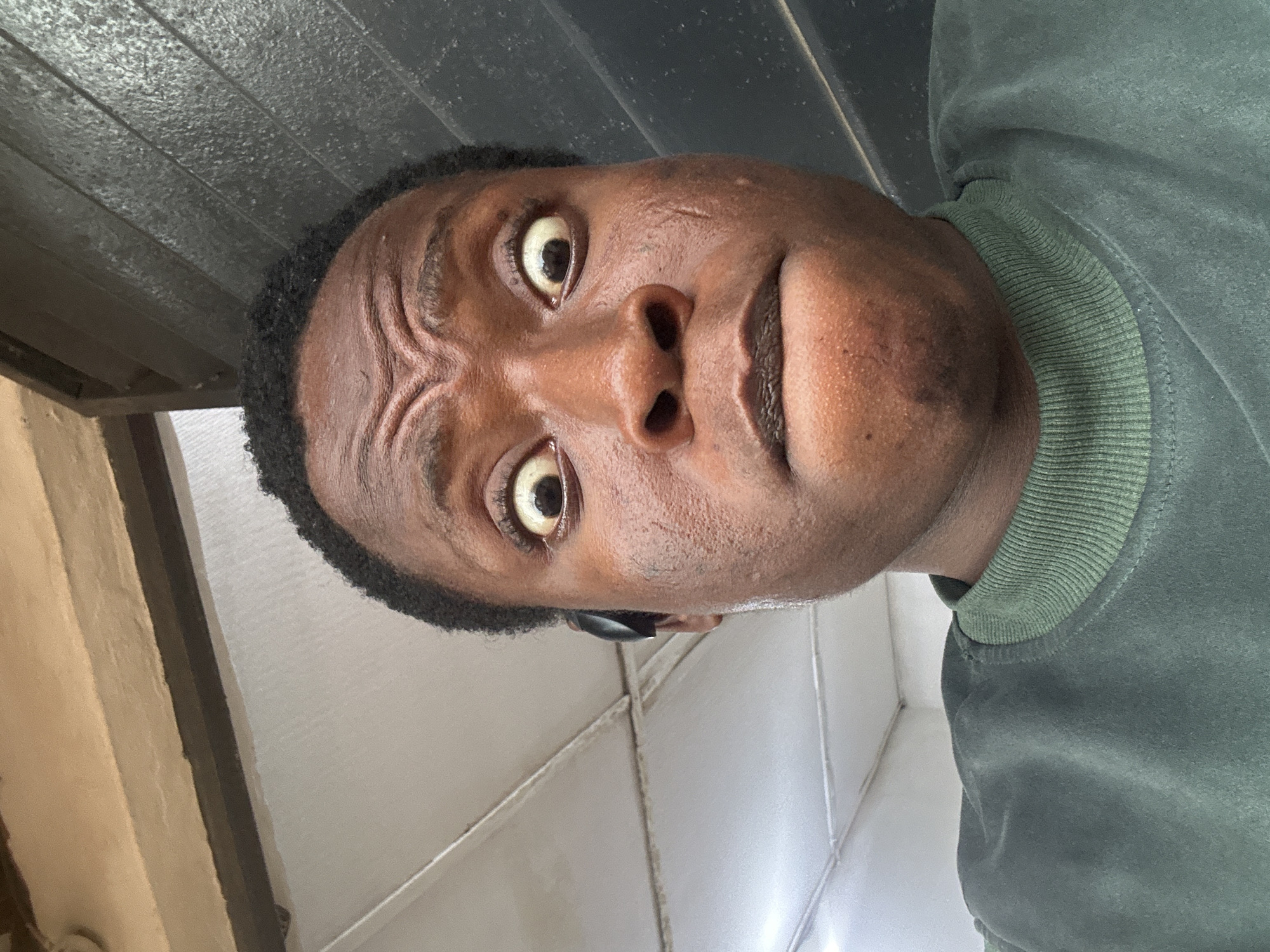 A close-up, low-angle, rotated view of a dark-skinned individual with tightly coiled hair, wearing a green sweatshirt. The person's face is prominent, featuring wide eyes, a wrinkled forehead, and visible pores on the skin. A black object is visible near the left ear. The background includes an undetermined dark, textured surface above and a combination of white and tan surfaces below, possibly walls. The location is unknown.