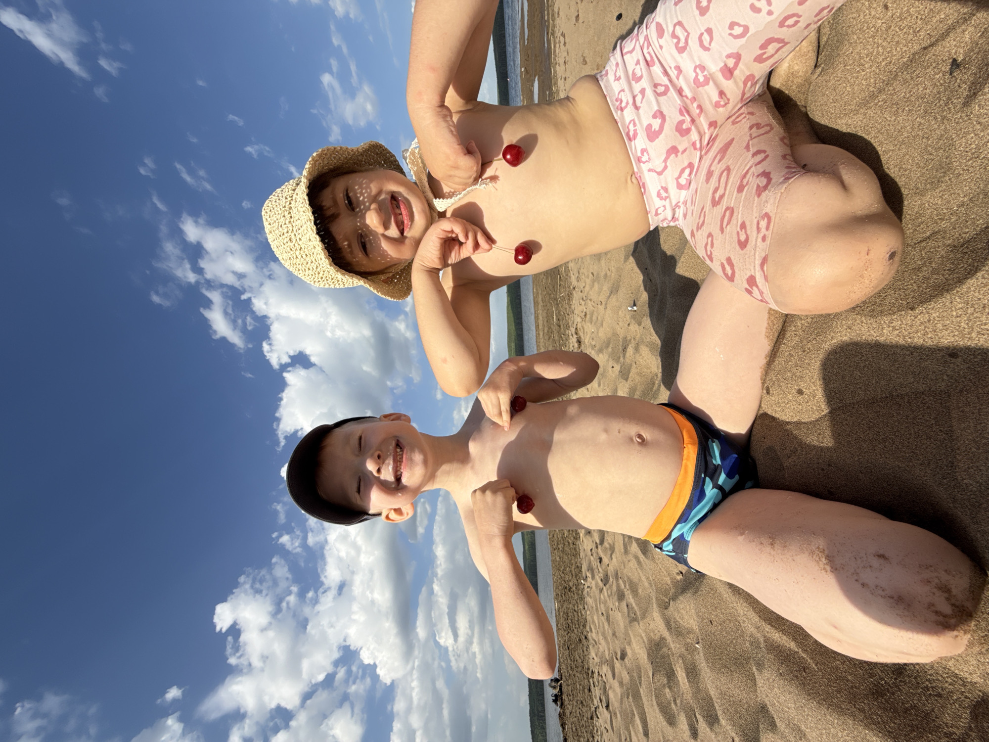 Two fair-skinned children, one wearing a straw hat and pink patterned leggings and the other a black cap and blue and orange swim trunks, are lying on a sandy beach at an angle to the camera. Each child is holding two cherries against their torso. The background shows a body of water and shoreline, and a sky with blue and white clouds. The location is Chaikovsky, Russia.