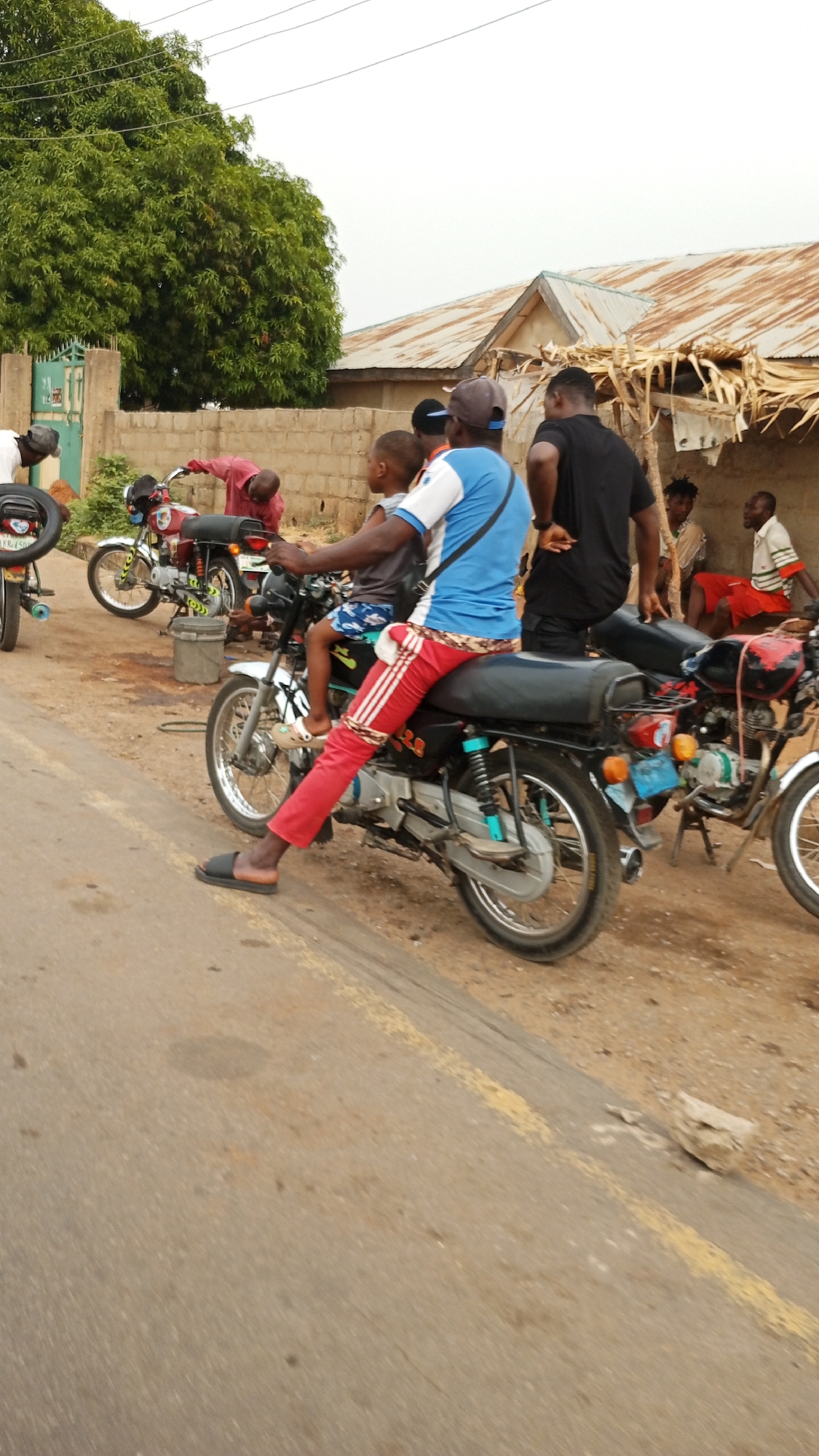 A man in red pants and a blue and white shirt sits on a black motorcycle, with a child in front of him. Several people are standing around motorcycles. A building with a corrugated metal roof sits in the background, with several people sitting under it. The location is along a paved road, likely in the city of Zango-ndaji, Nigeria.