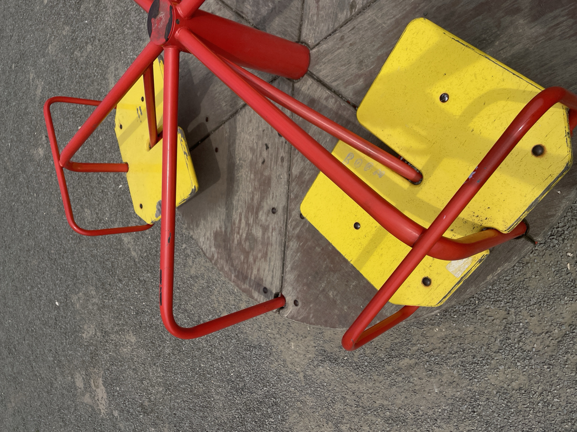 The image shows a red and yellow merry-go-round on a gravel surface, likely in a park or playground. The merry-go-round has four yellow seats with red metal bars for support and seating. The wooden base of the merry-go-round is visible, showing signs of wear and tear. The gravel ground is dark gray and appears to be a common surfacing material for playgrounds. No people are present in the image, and there are no indications of any specific location within Rostov-on-Don, Russia. The overall scene depicts a simple, functional piece of playground equipment in an outdoor setting.