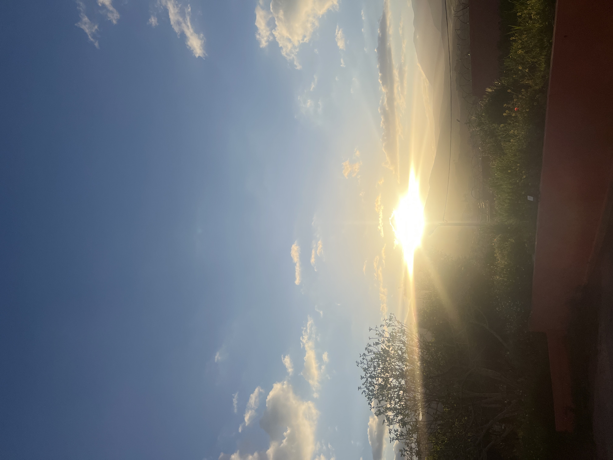 Here's a detailed description of the image:
**General Impression:**
The image shows a sunlit scene with a view of a bright sky, clouds, and part of a landscape. The strong sunlight makes it difficult to discern specific details.
**Sky:**
The sky is a mix of blue and light white/yellow due to the sun. There are scattered clouds.
**Landscape:**
The right side of the image features foliage and some structure, potentially a building, partially obscured by the bright sunlight. Some hills and a town are also visible, obscured by sunlight. The photo was taken in Ouaouizert, Morocco.
